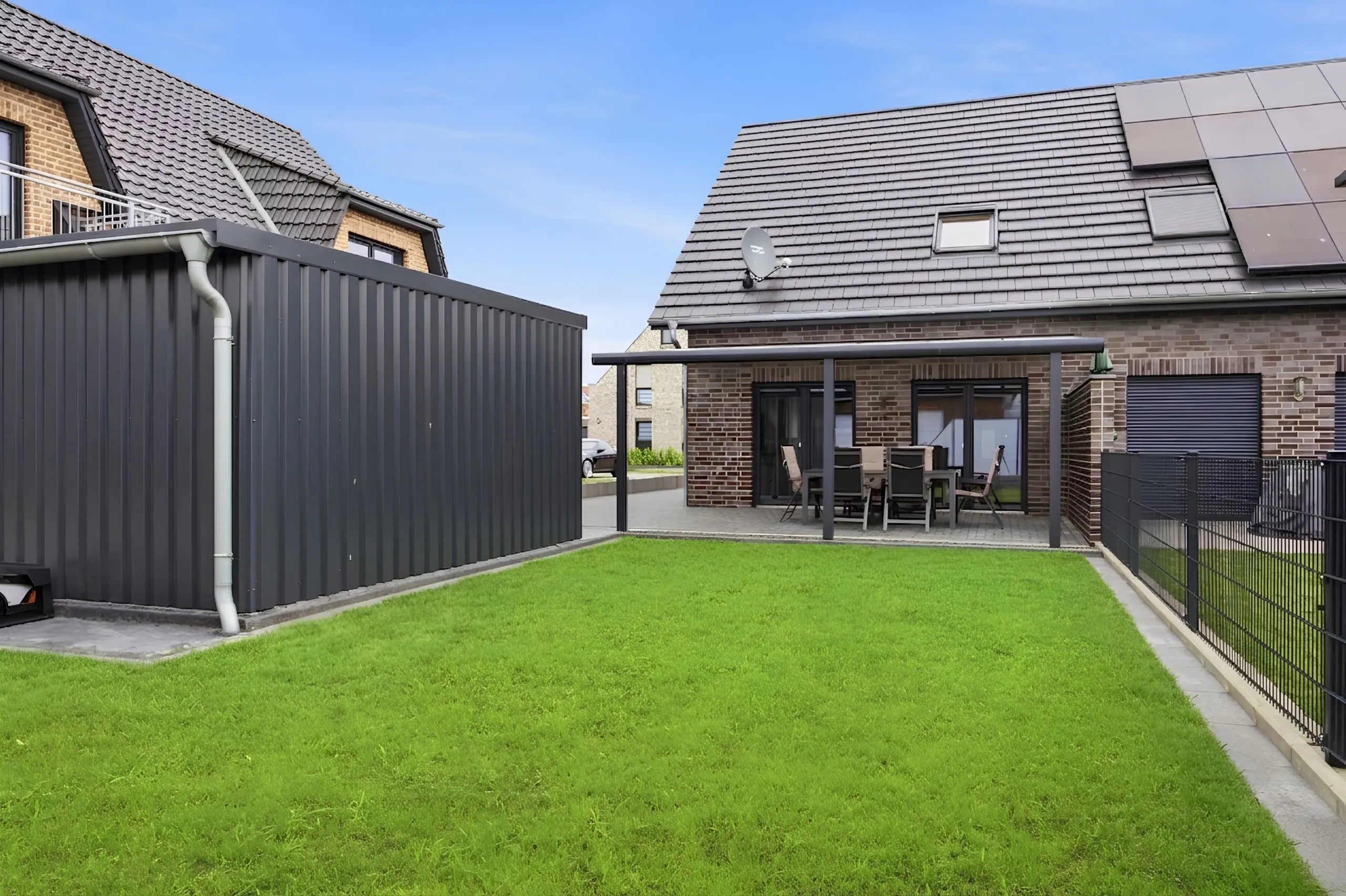 Backyard of a modern brick house with a dark metal fence, patio table and chairs, and a satellite dish on the roof.