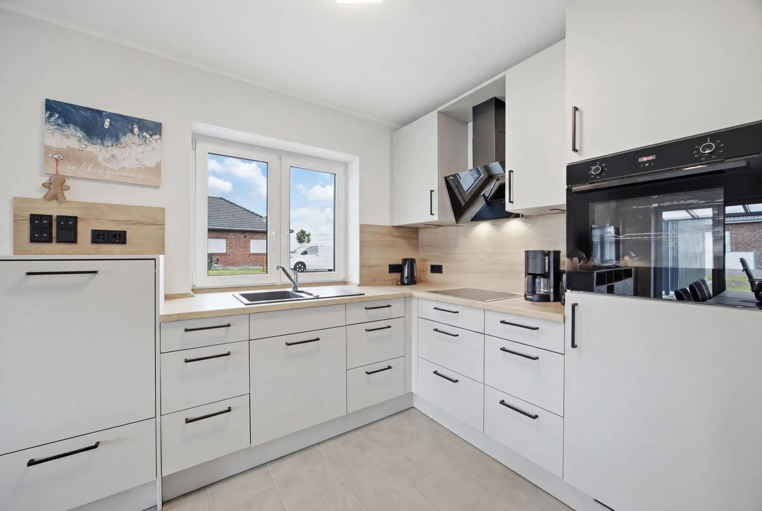 Bright modern kitchen with white cabinets, beige countertops, and a large window over the sink beside a built-in oven suite on the right side.