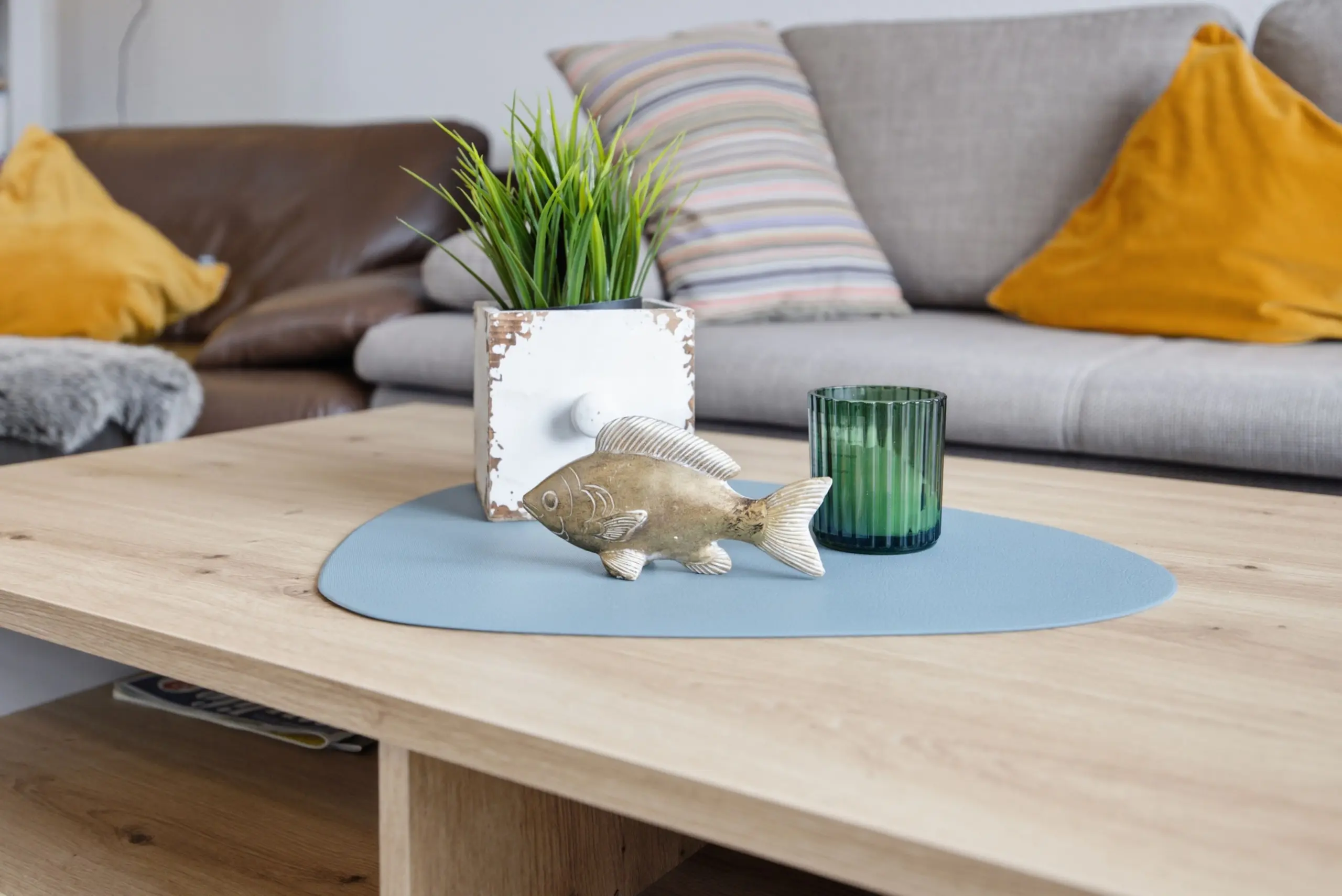Living room coffee table with a spiky green plant in a white distressed pot, a blue placemat, a green ribbed glass, and a decorative fish statue; sofa with yellow cushions in background.