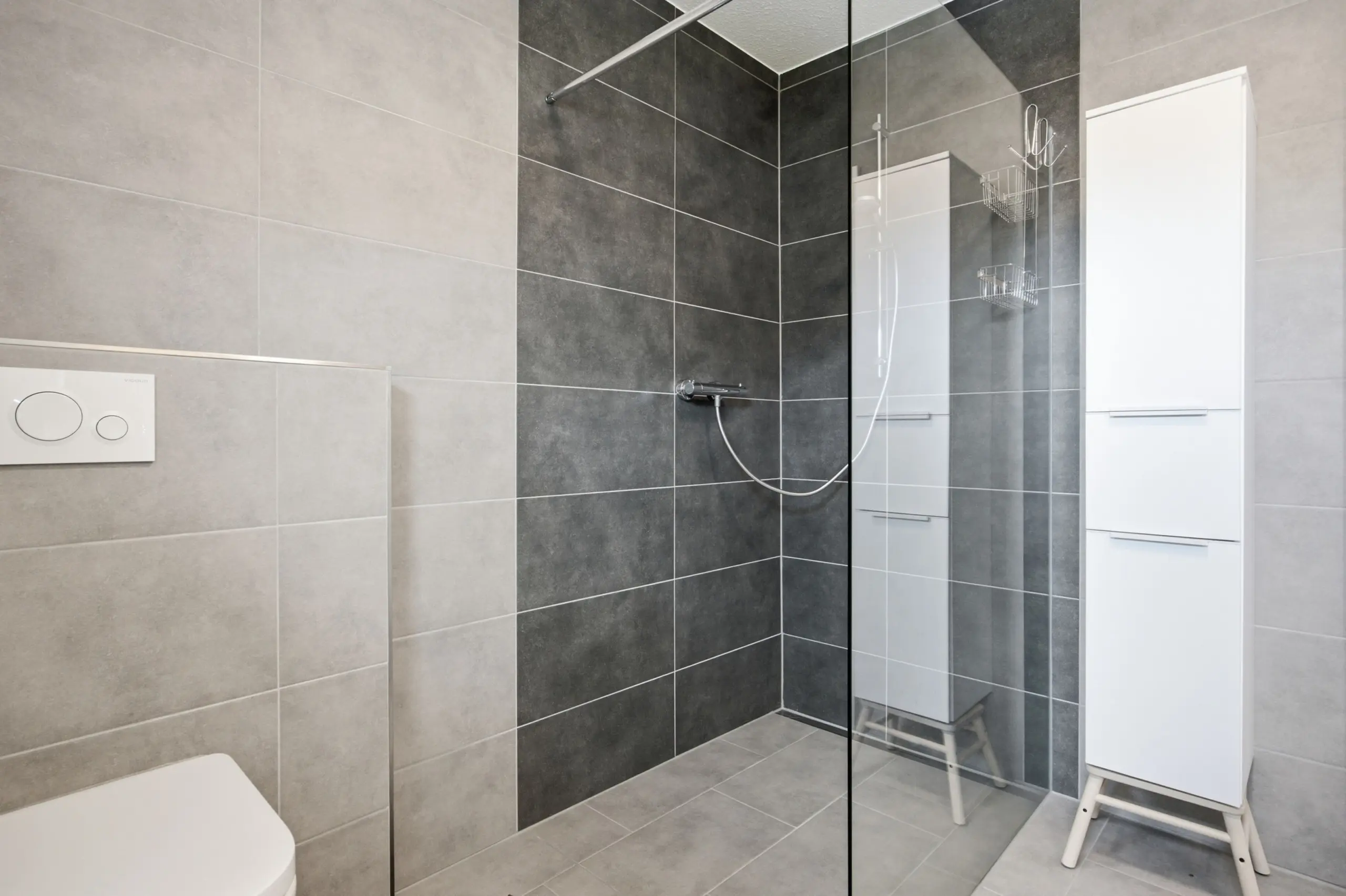 Modern bathroom with gray tiled walls and floors, a glass shower divider, a white vanity cabinet, and a partial toilet visible.