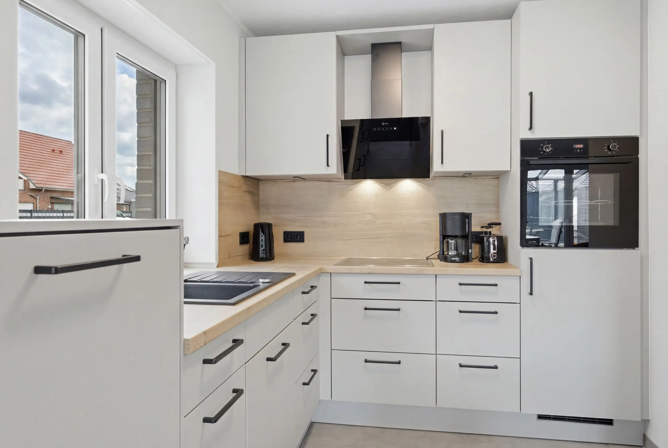 Modern white kitchen with a stainless steel range hood above a black oven and light wood countertops.