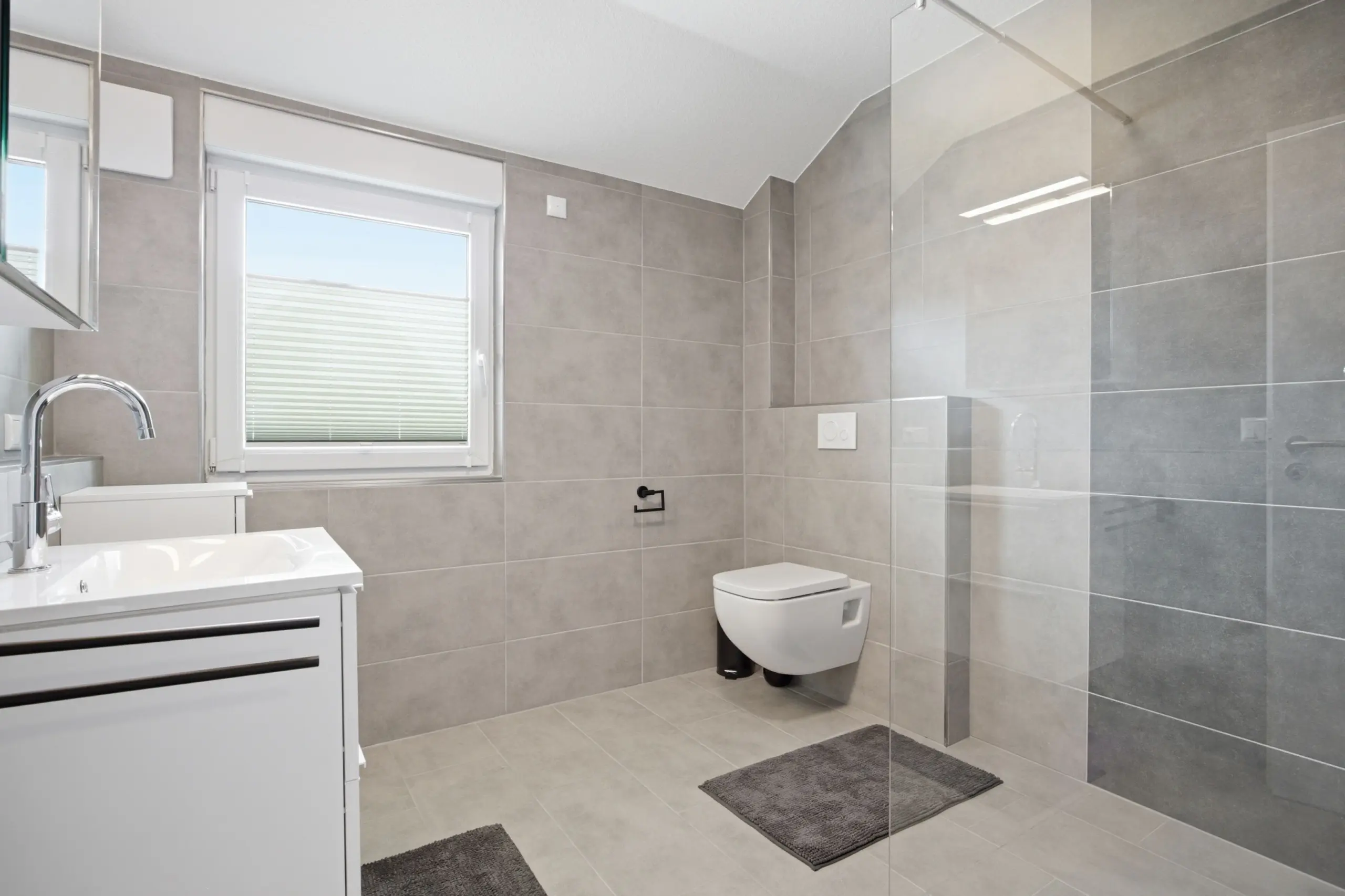 Modern gray-tiled bathroom with a wall-mounted toilet, white vanity, and a glass shower enclosure.