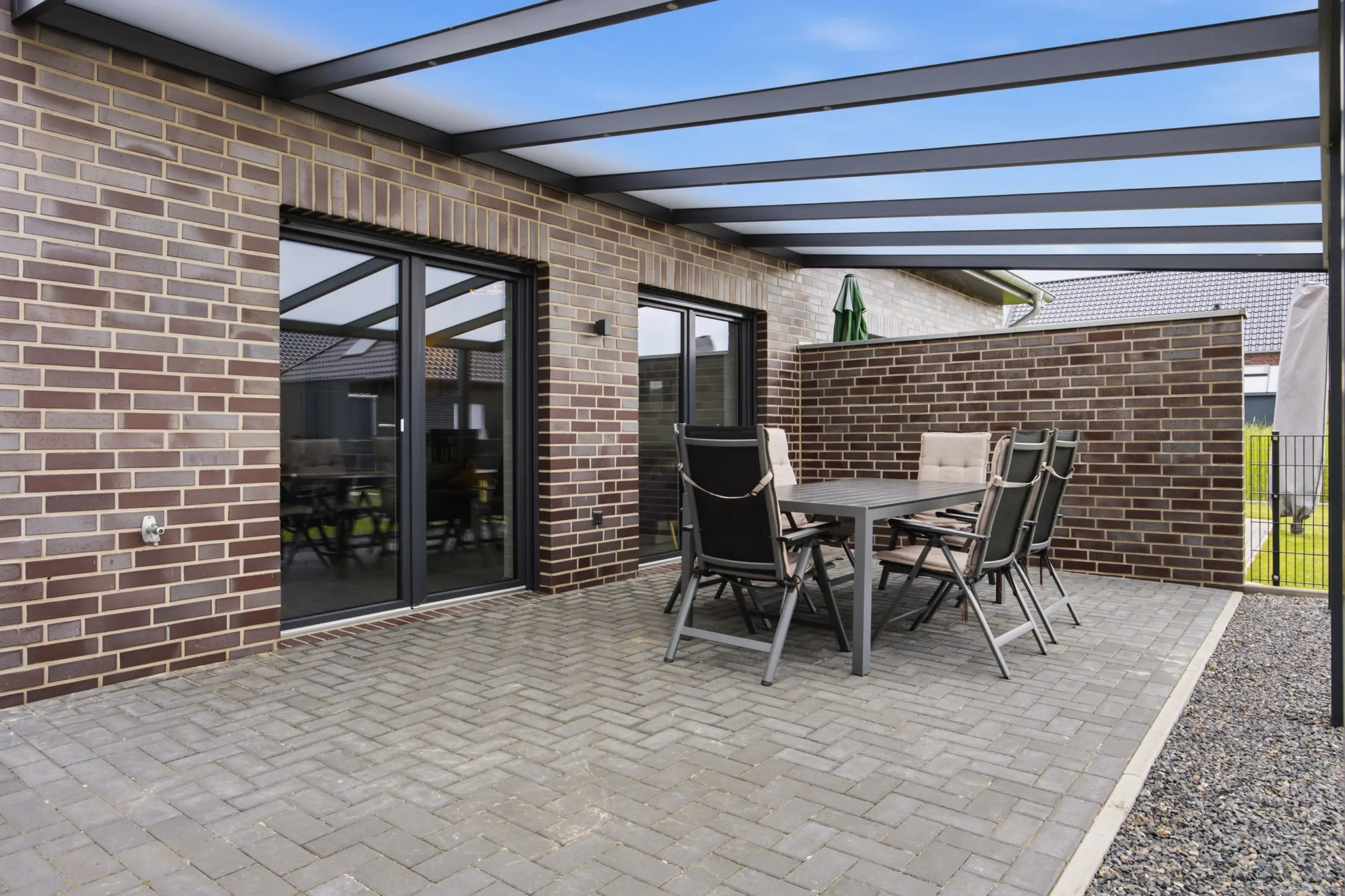 Patio area of a brick house with sliding glass doors, dark metal pergola beams, and a gray outdoor dining table with six chairs and a folded green umbrella on the side.",