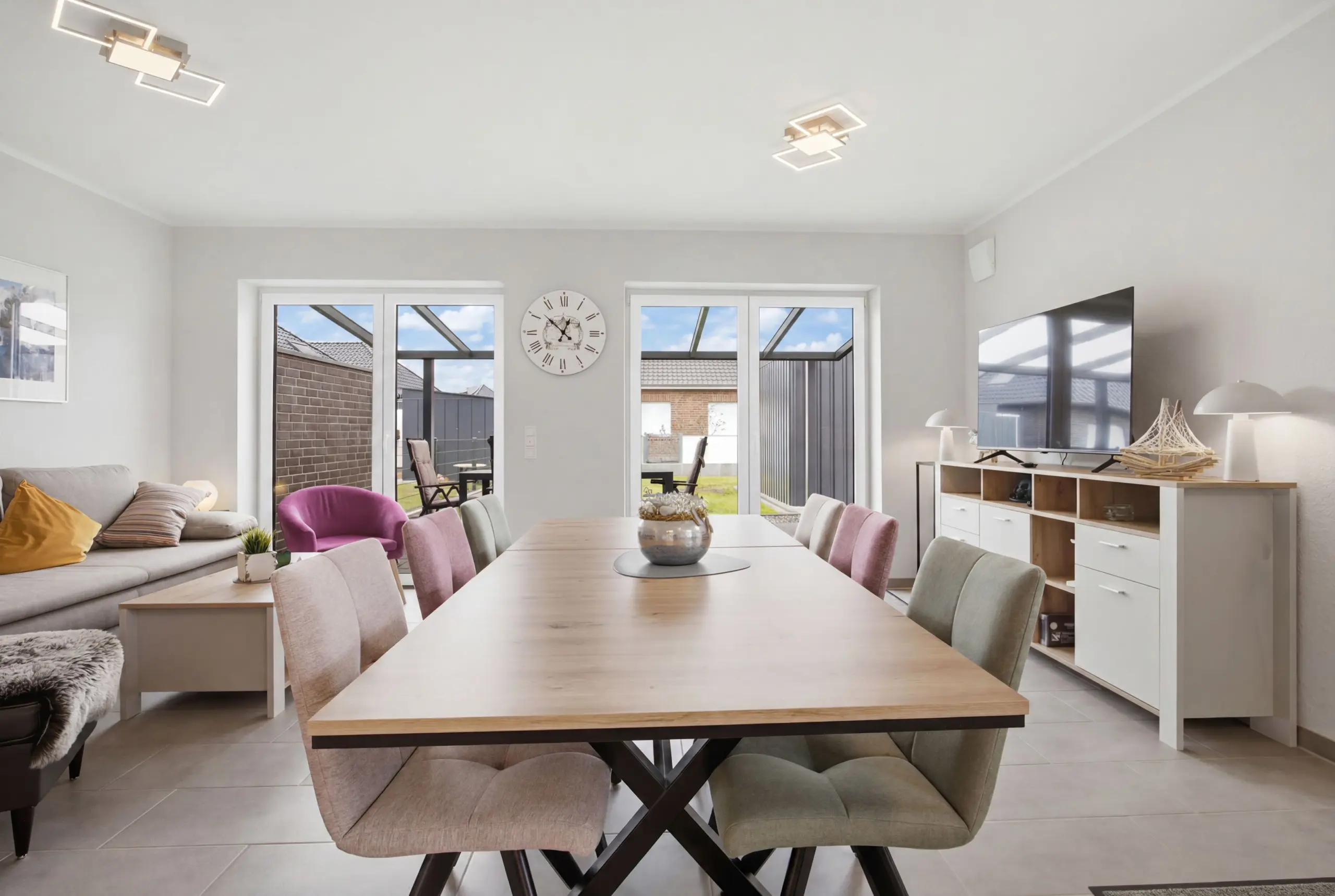 Open-plan living and dining room with a long light wood table, pink/neutral chairs, a wall clock between glass doors, and a TV along a side cabinet.