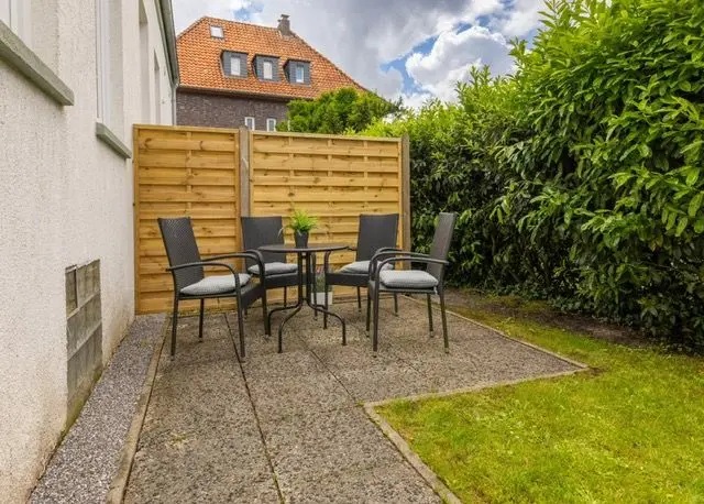 Outdoor patio with a round table and five metal chairs on a paved area, wooden privacy fence behind, and lush hedges to the right.