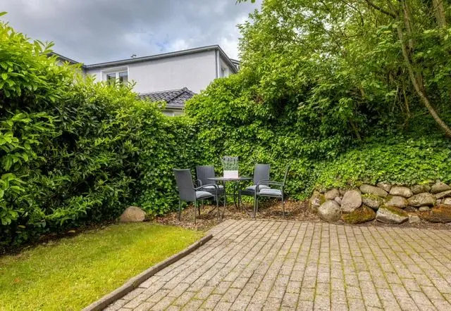 Patio dining set in a green backyard: round glass table with four gray chairs, surrounded by hedges and a stone border across a paved area.