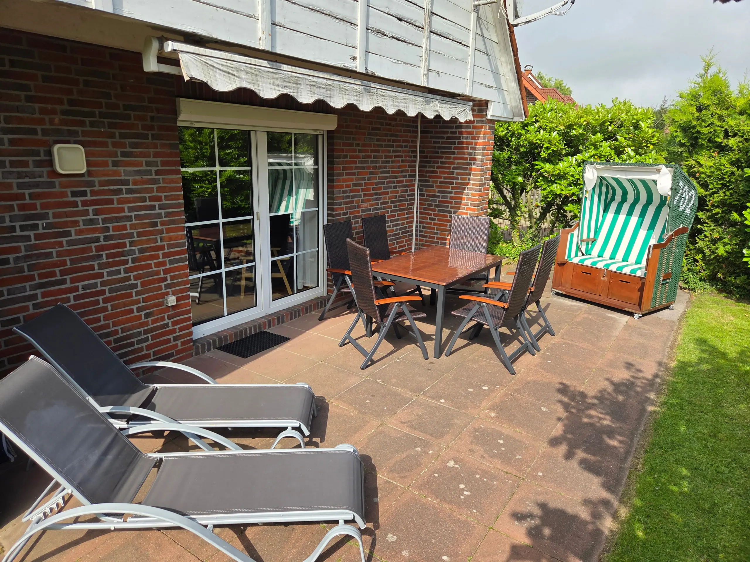 Backyard patio with brick house, glass door, a wooden dining table and wicker chairs under a white awning. Back right shows a striped green-and-white beach chair and green hedges.