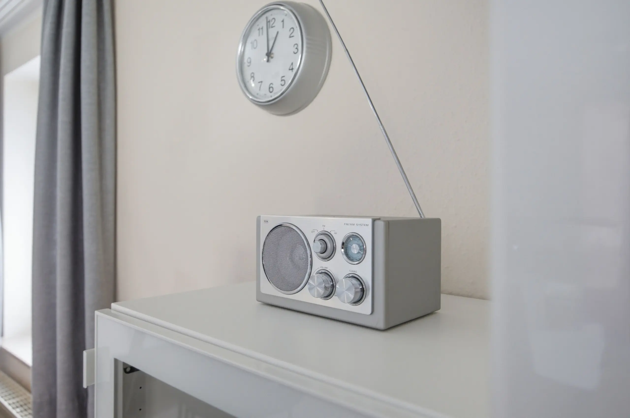 Grey vintage radio with dials rests on a white surface beneath a round wall clock in a bright room.
