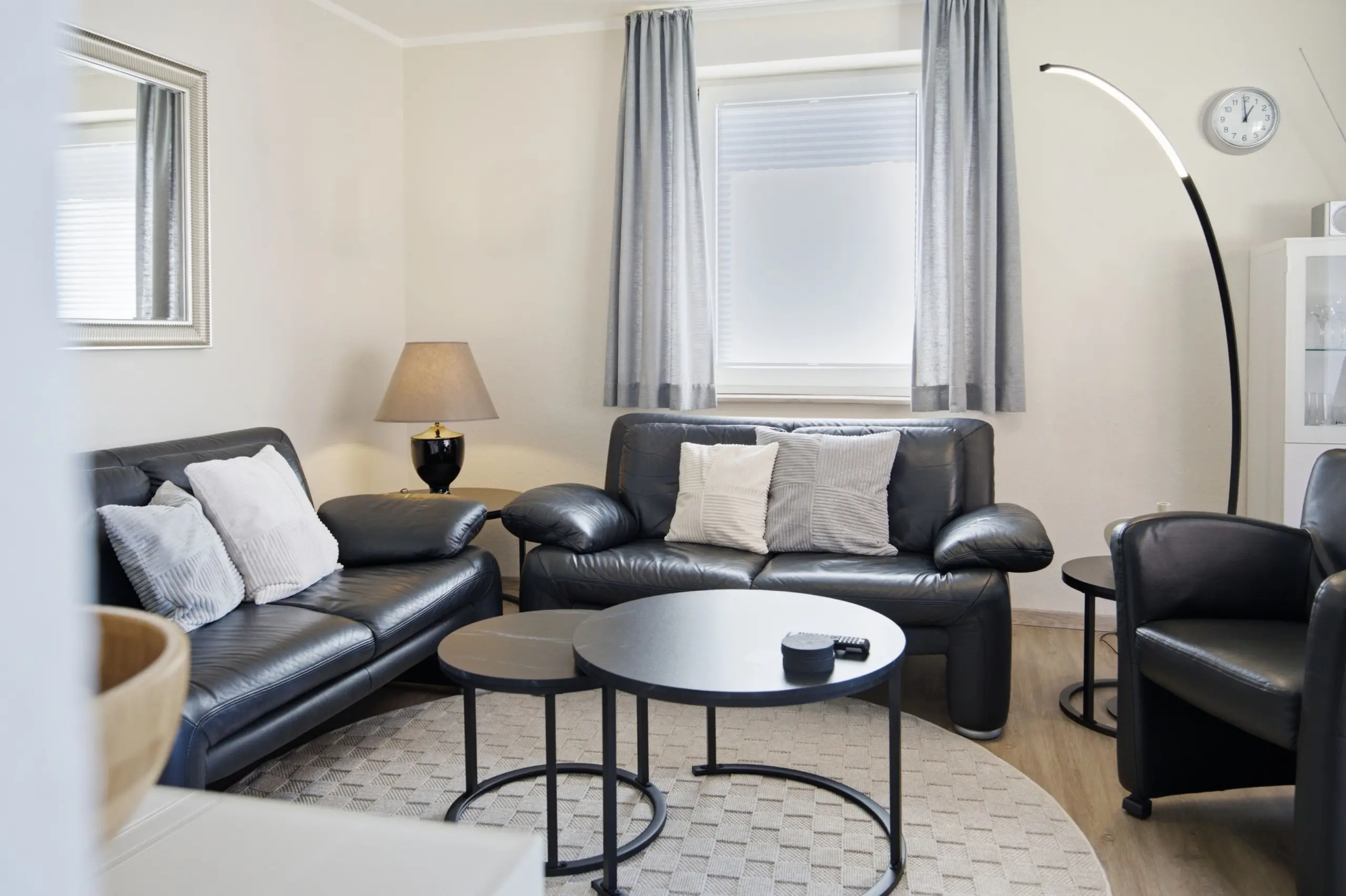 Living room with black leather sofa set and gray throw pillows, two round coffee tables, and a tall arc floor lamp near a window with curtains.