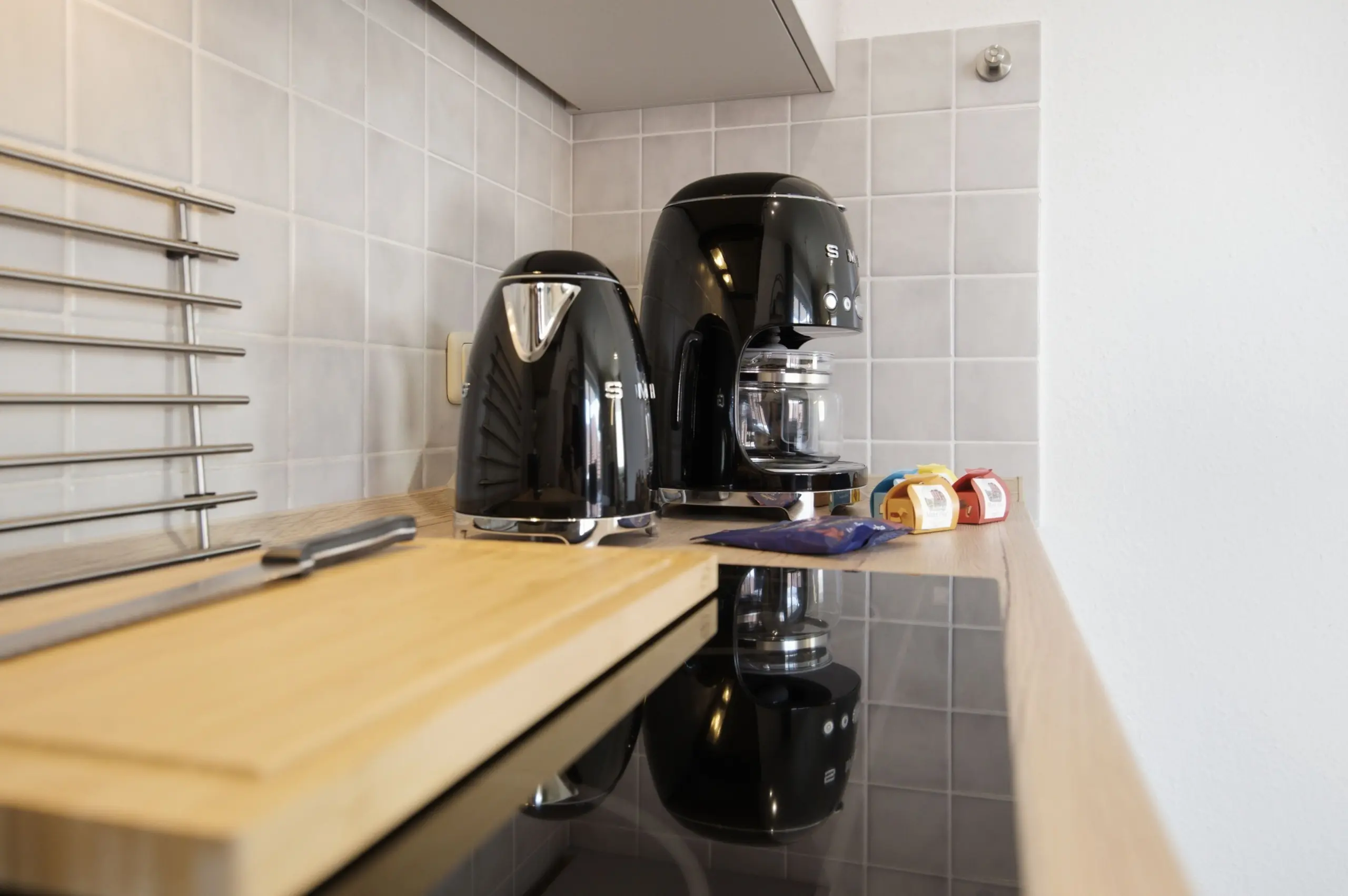 Two black coffee machines sit on a light wooden kitchen counter against a tiled backsplash, with colorful coffee pods nearby and a metal rack on the left.