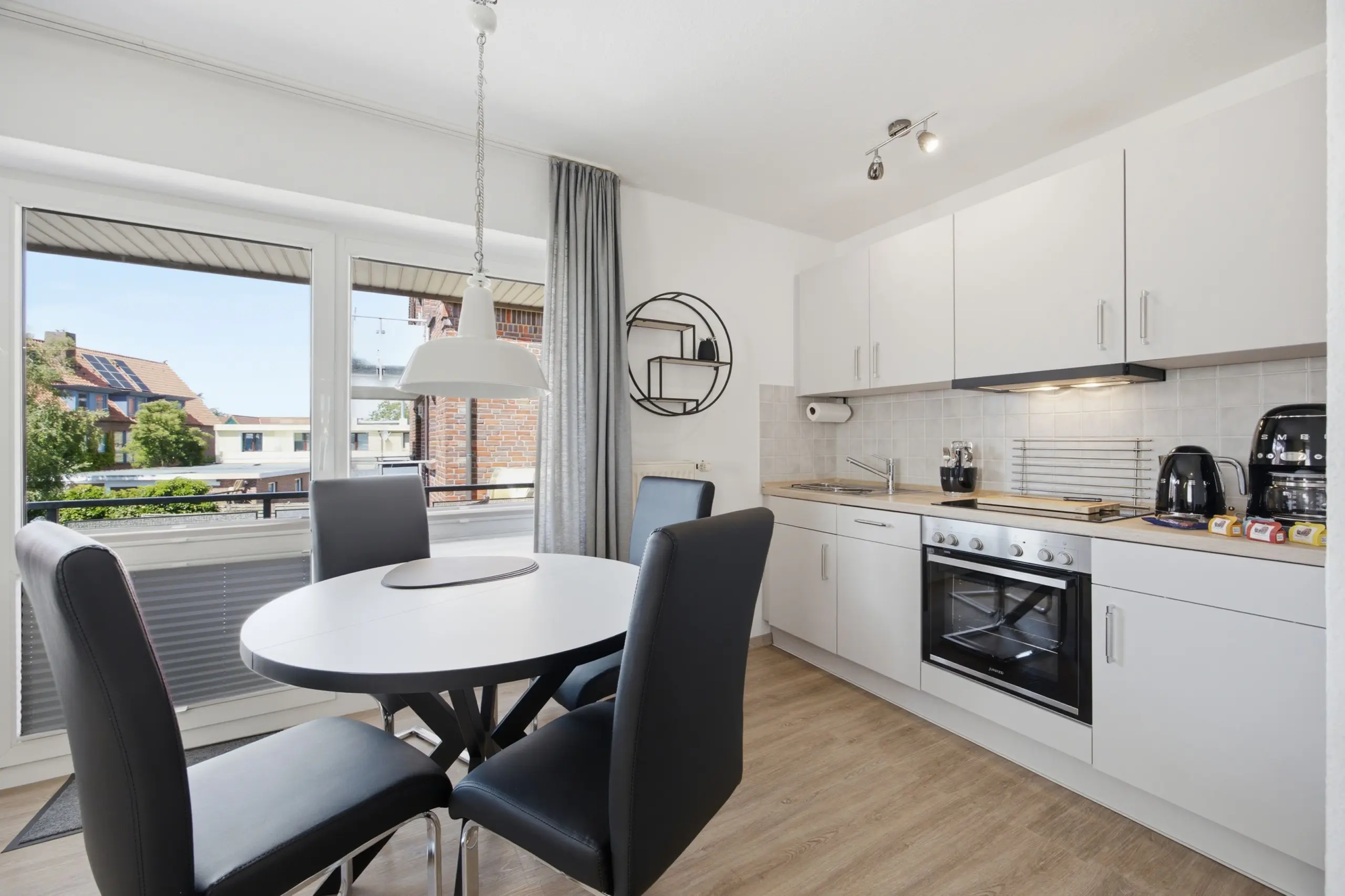 Bright kitchen and dining area with white cabinets, stainless steel oven, and a round table by a large window showing buildings outside.