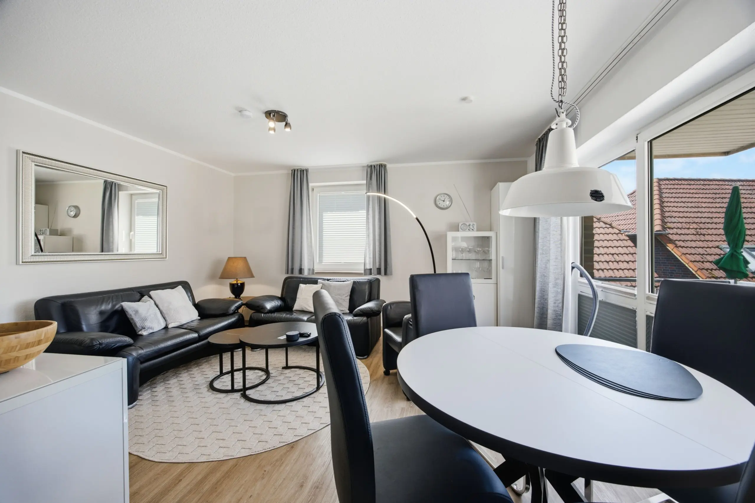 Open-plan living and dining room with black leather sofas, a glass coffee table, and a large mirrored wall reflecting light from windows behind gray curtains.