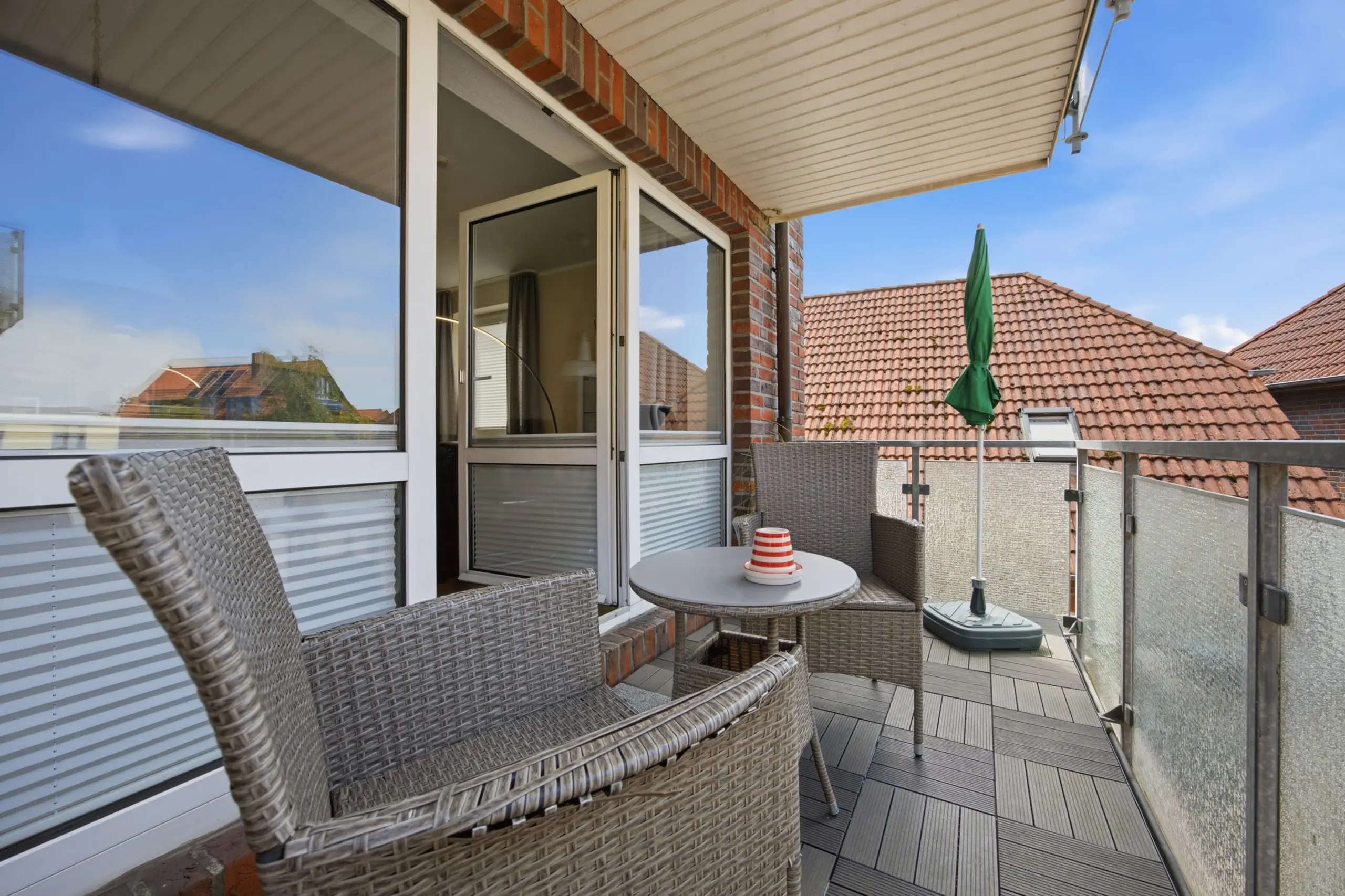 Brick apartment balcony with wicker chairs, glass railing, and a closed green umbrella against a blue sky.