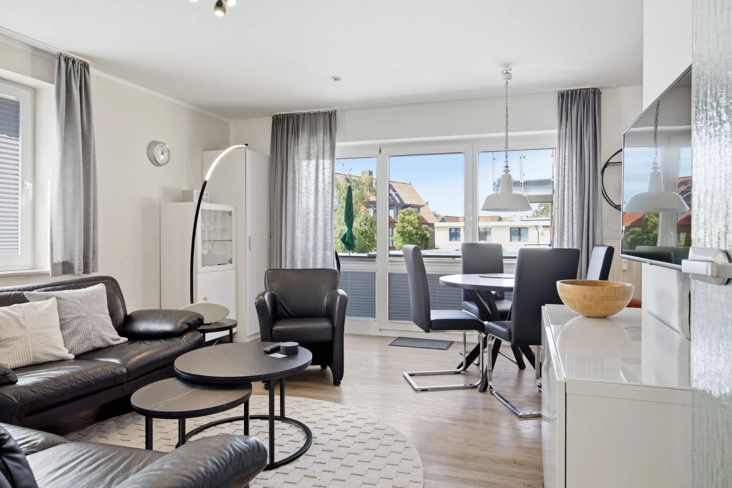 Modern open-plan living and dining area with gray curtains, black leather seating, and round black coffee tables by large windows.