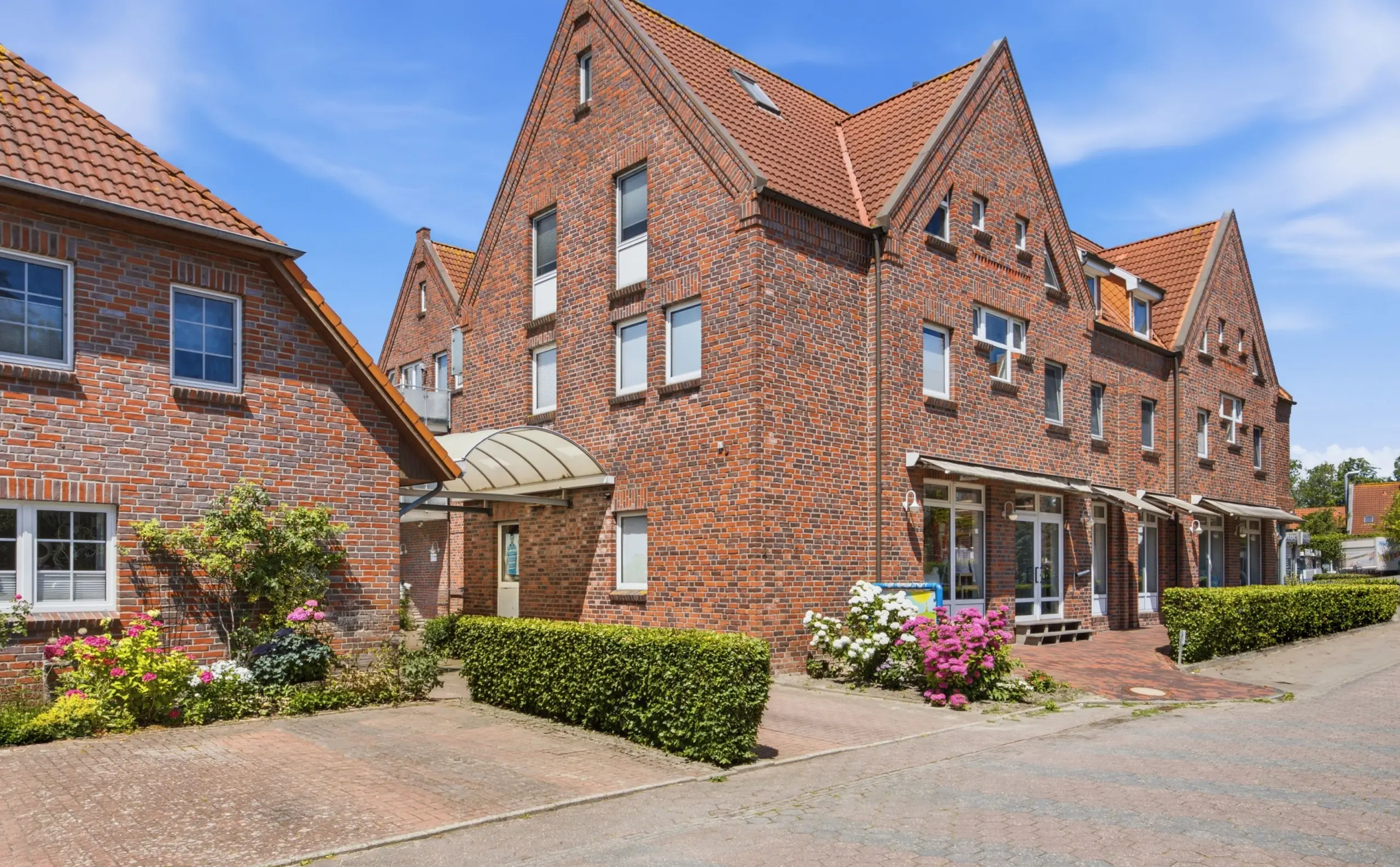 Brick residential building with stepped gables and orange tiled roofs under a clear blue sky, flowering shrubs in front.
