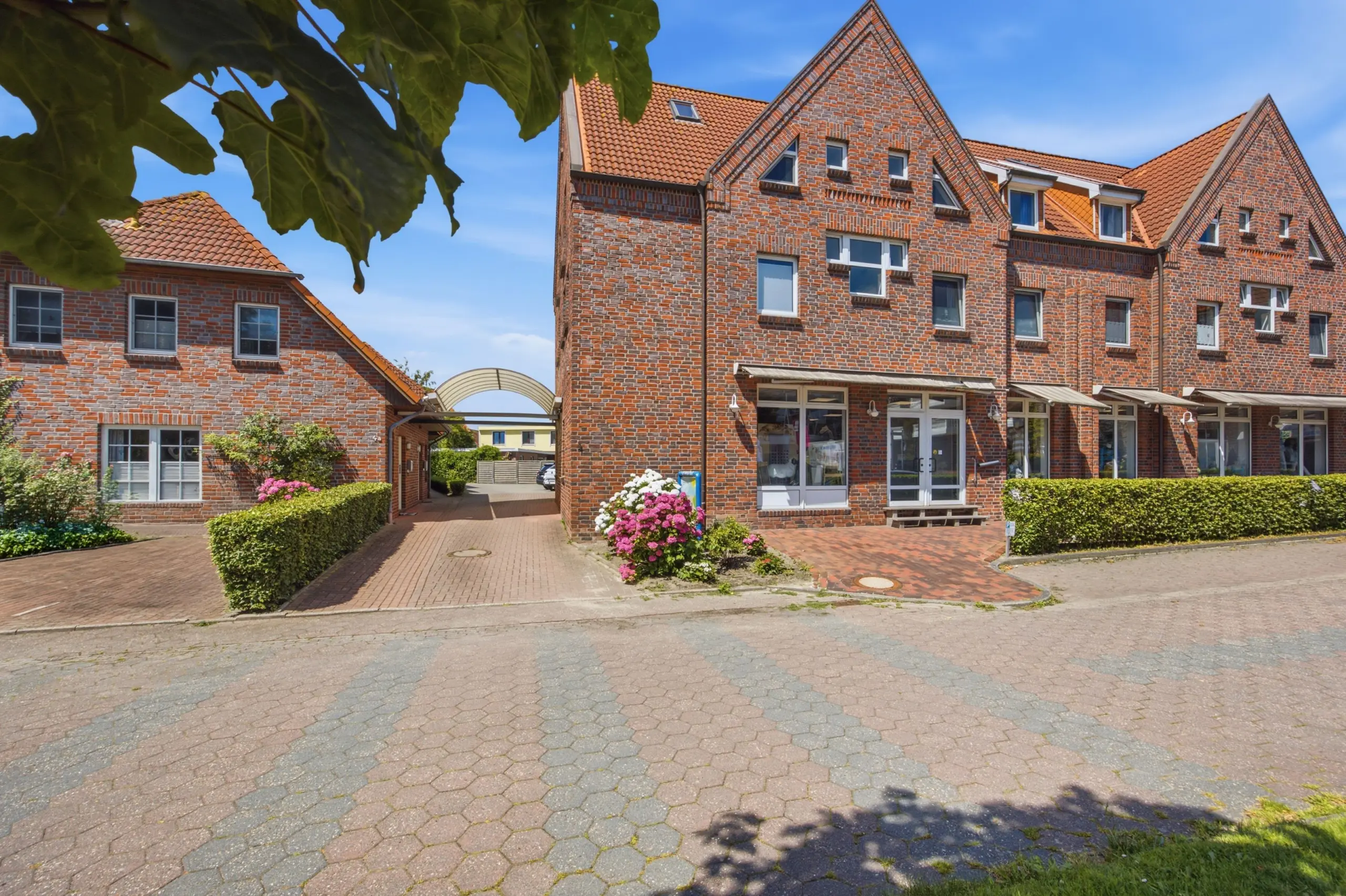 Brick row houses with orange-tiled roofs lining a pedestrian street under a blue sky; flowering bushes and hedges in front.