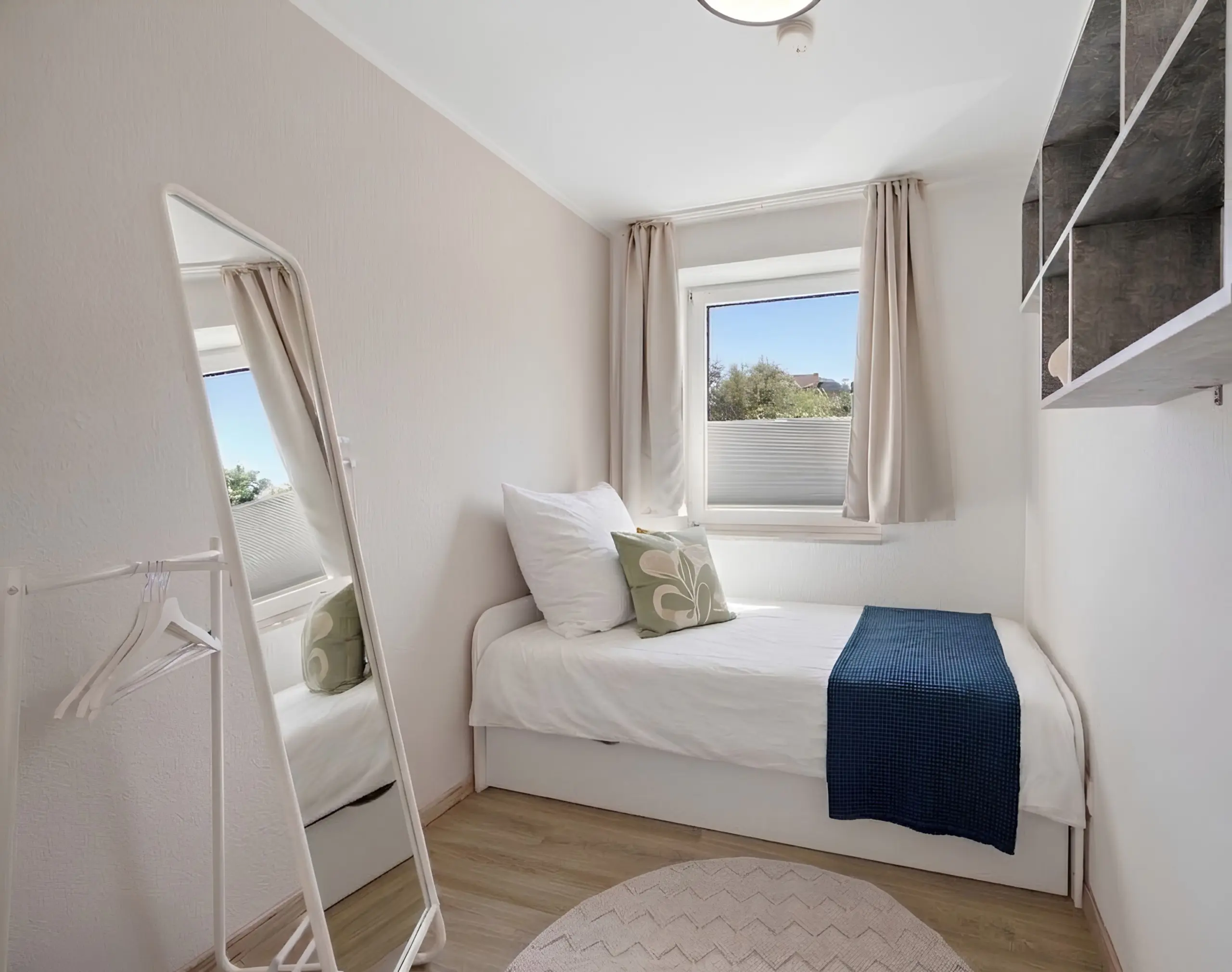 Small bright bedroom with a white bed, blue throw, and a window with beige curtains; a leaning mirror is on the wall.