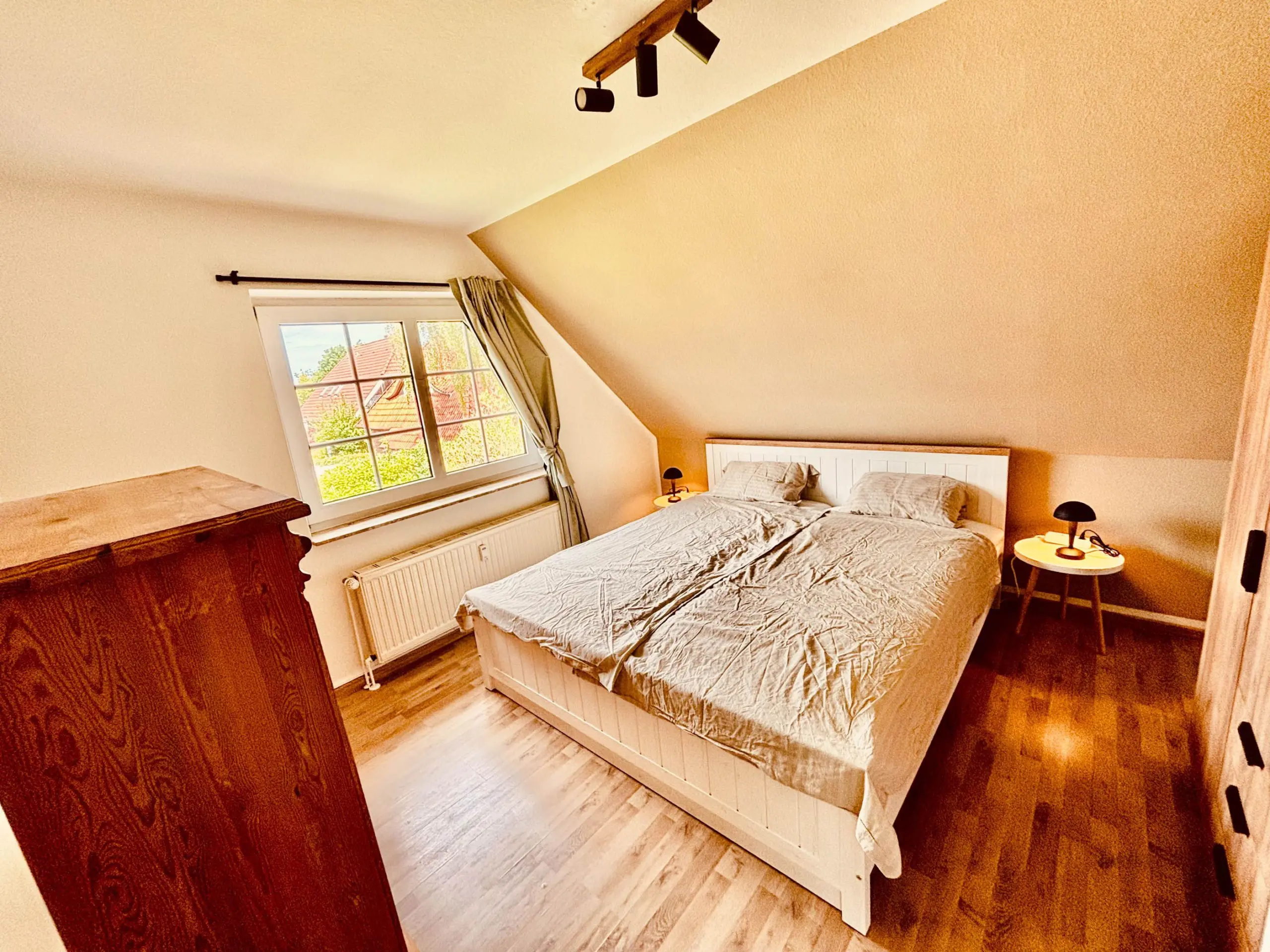 Bright bedroom with two single beds, beige bedding and white headboards, plus two round nightstands and a large window overlooking a sunny yard.