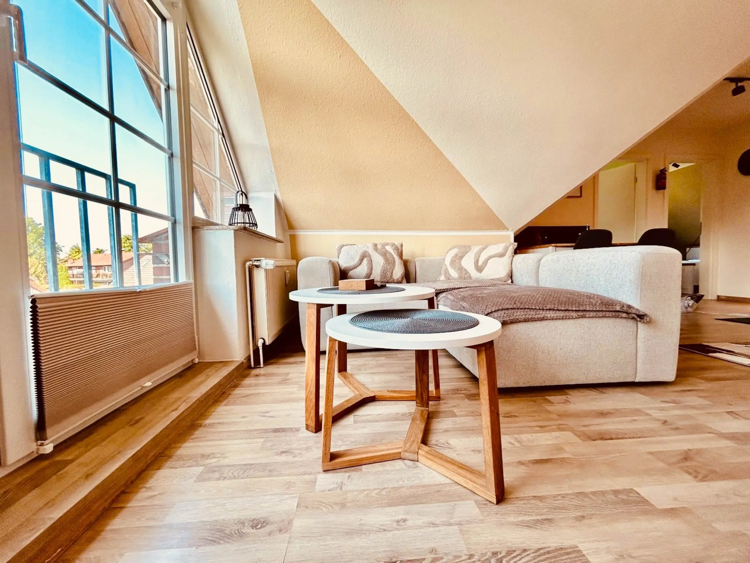 Bright living room with a large angled ceiling, big windows, and a beige sofa with patterned cushions facing two round nesting tables.