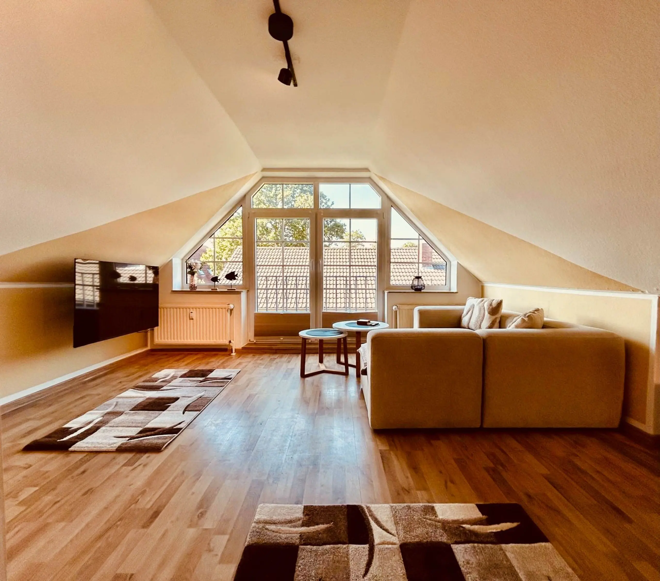 Living room with a large triangular window, sloped ceiling, and beige furniture; sunlight fills the space via glass doors and windows.