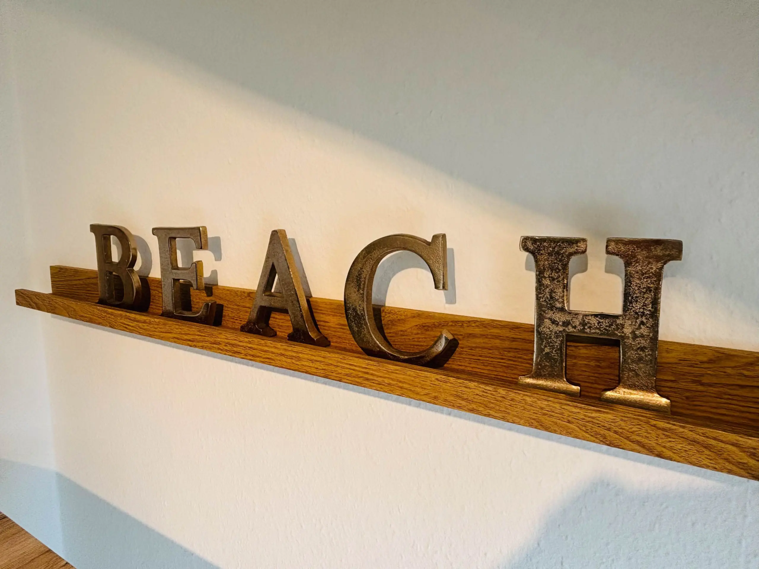 Decorative metal letters spelling 'BEACH' on a wooden wall shelf against a light wall.