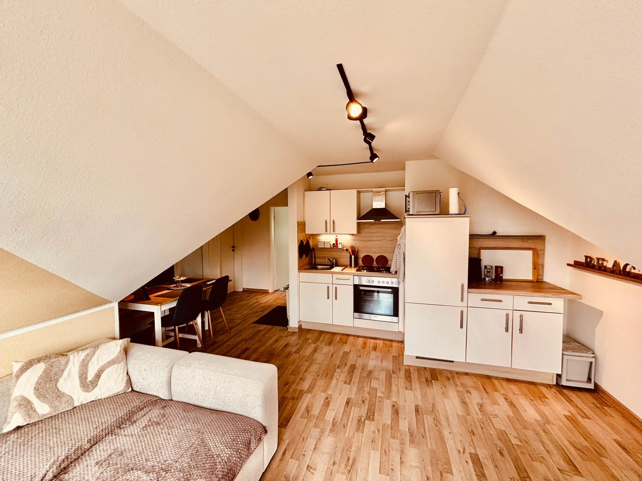 Open-plan attic kitchen and living area with white cabinets, stainless oven, and a beige sofa in the foreground under a sloped ceiling with track lighting.