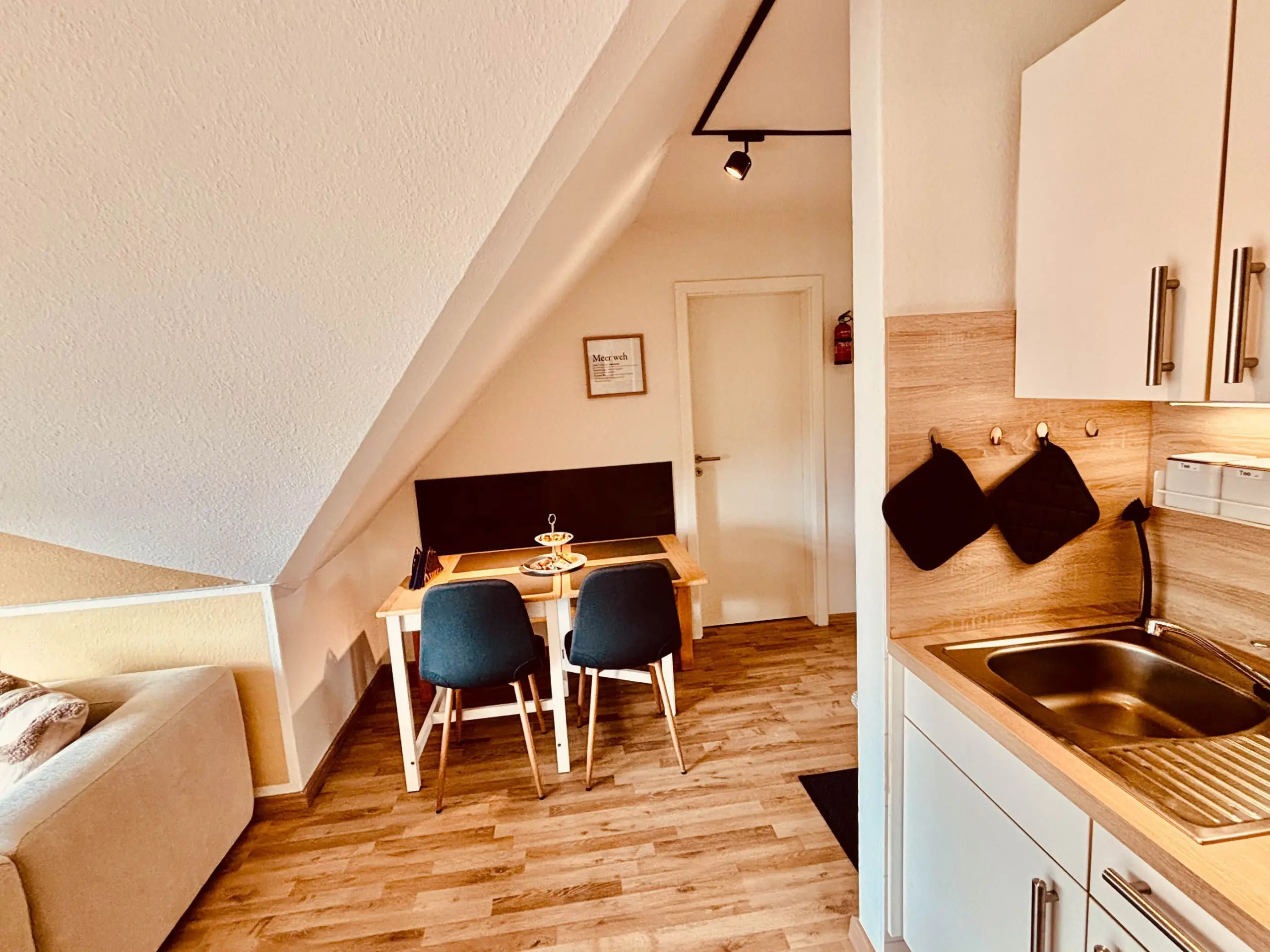 Attic dining area with a rectangular table set with plates, three blue chairs, a beige sofa on the left, and white kitchen cabinets on the right under a slanted ceiling.