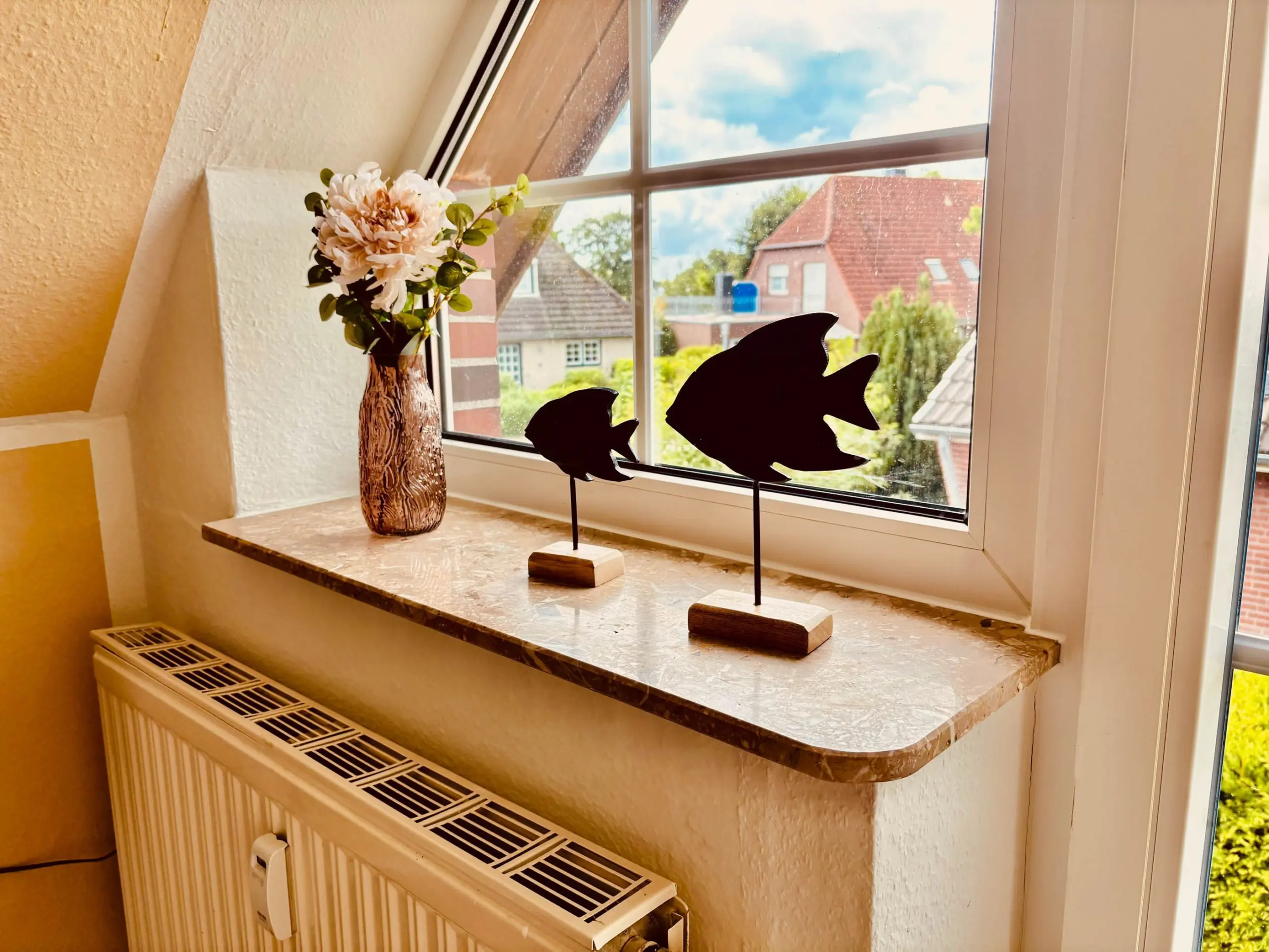 Vase with pale pink flowers on a marble windowsill, next to two black fish-shaped metal sculptures by a sunny window.