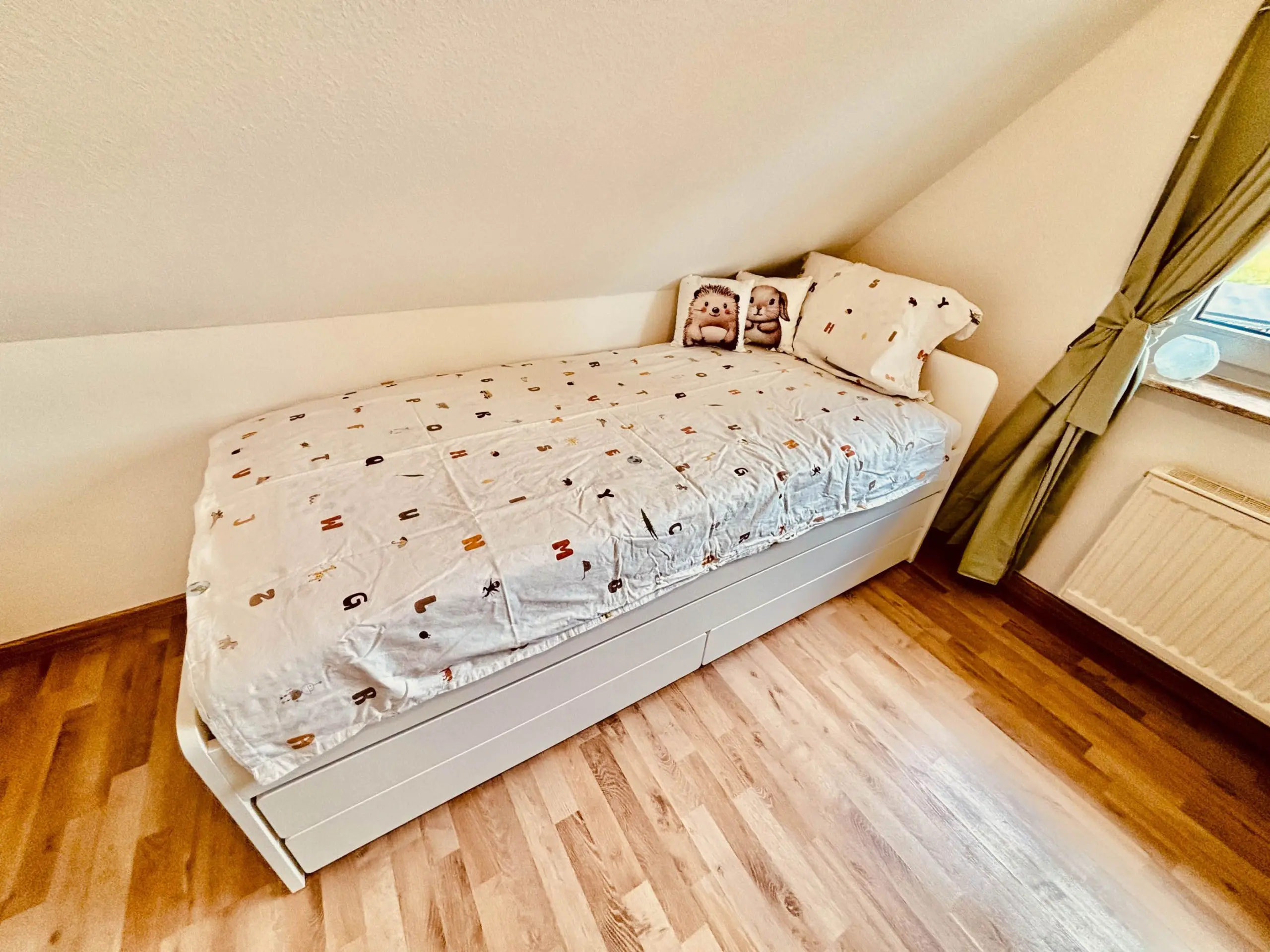 White child’s bed with an alphabet-pattern quilt and two animal-themed pillows under a sloped ceiling, beside a window with tied-back beige curtains.