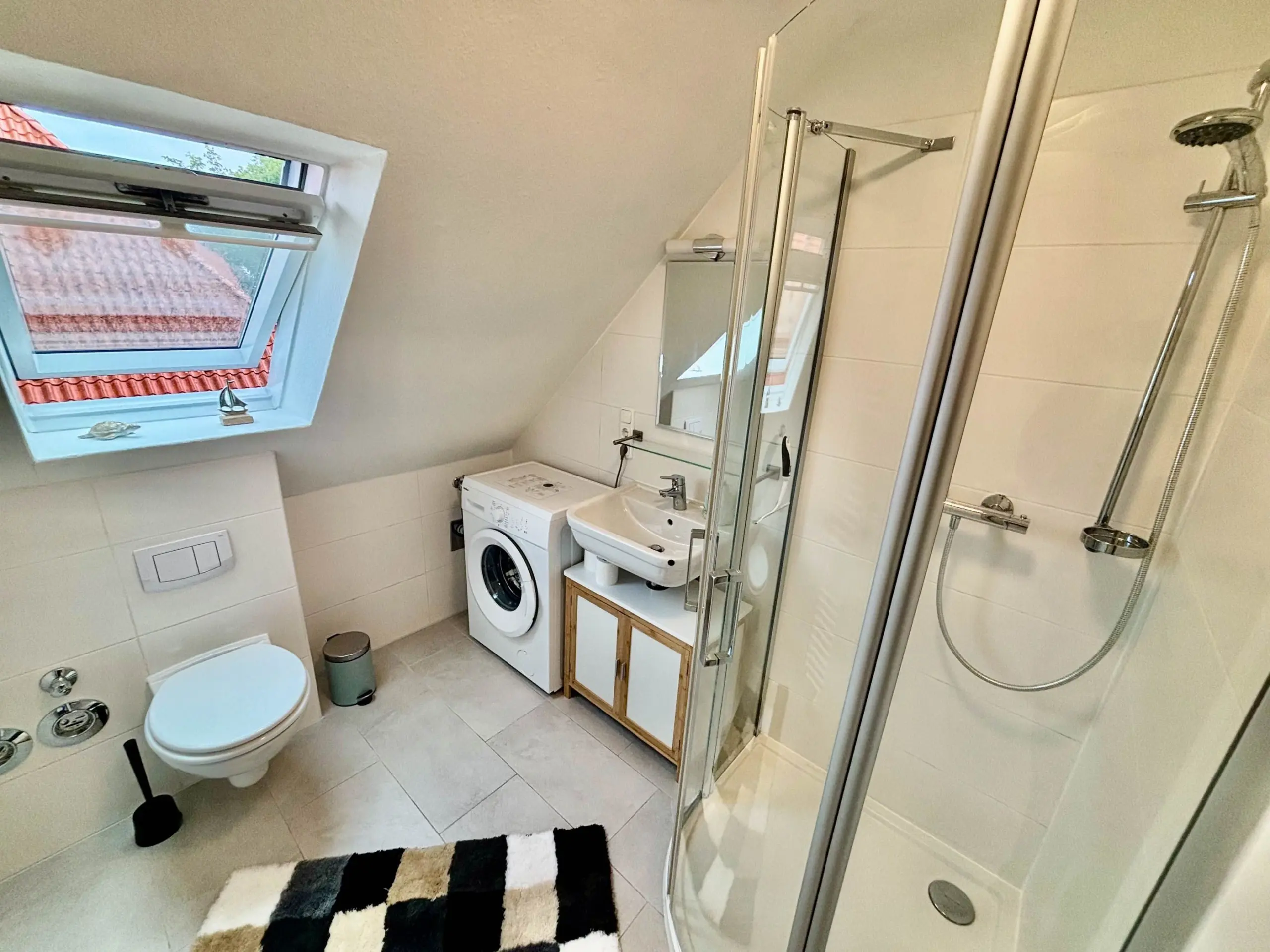 Small bright bathroom with a front-loading washing machine, sink, toilet, and glass shower enclosure; window shows red tiled roof outside.