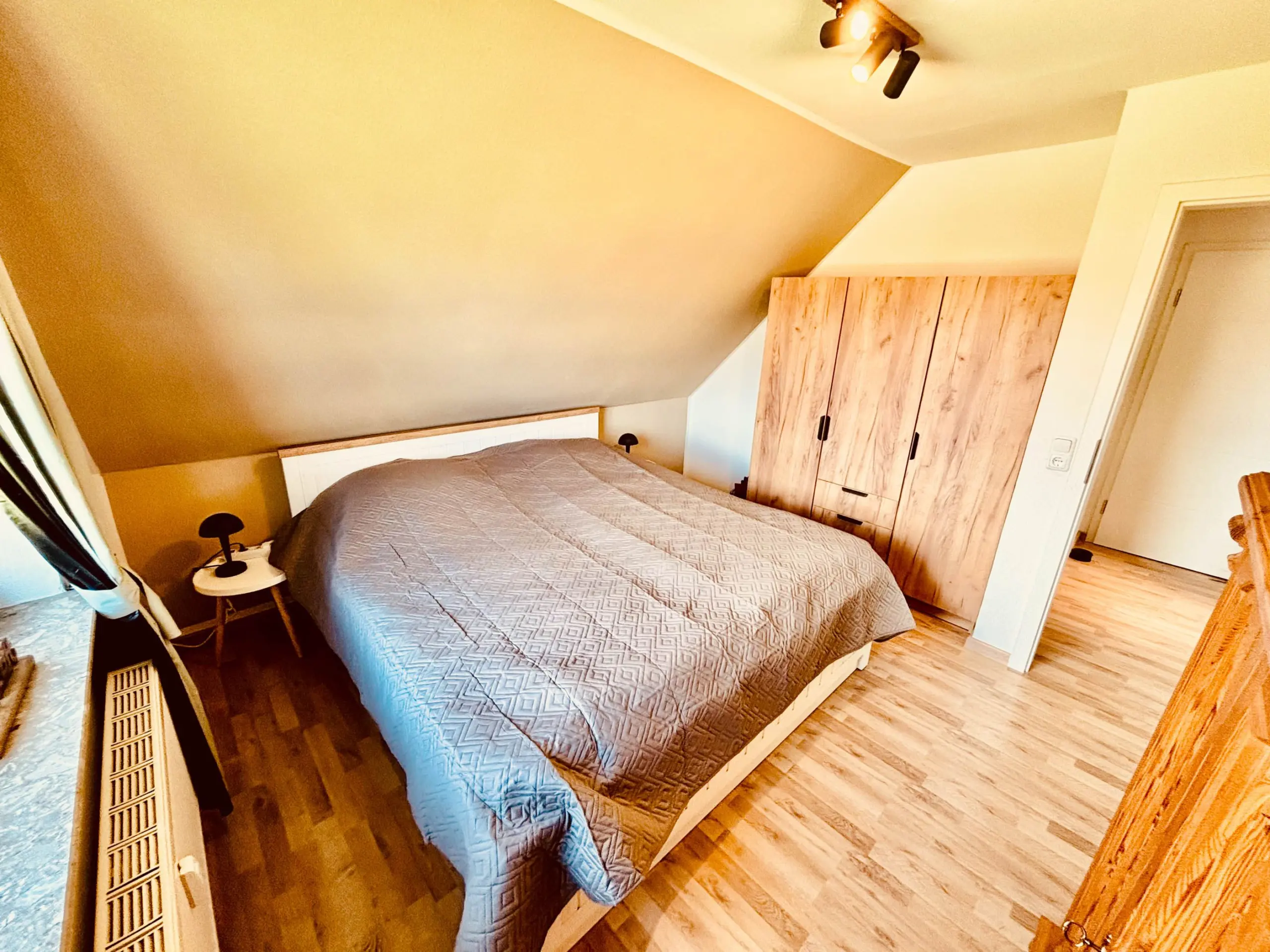 Angled-ceiling bedroom with a large wooden wardrobe, a bed in the foreground with a gray quilt, and a small round bedside table on the left side.