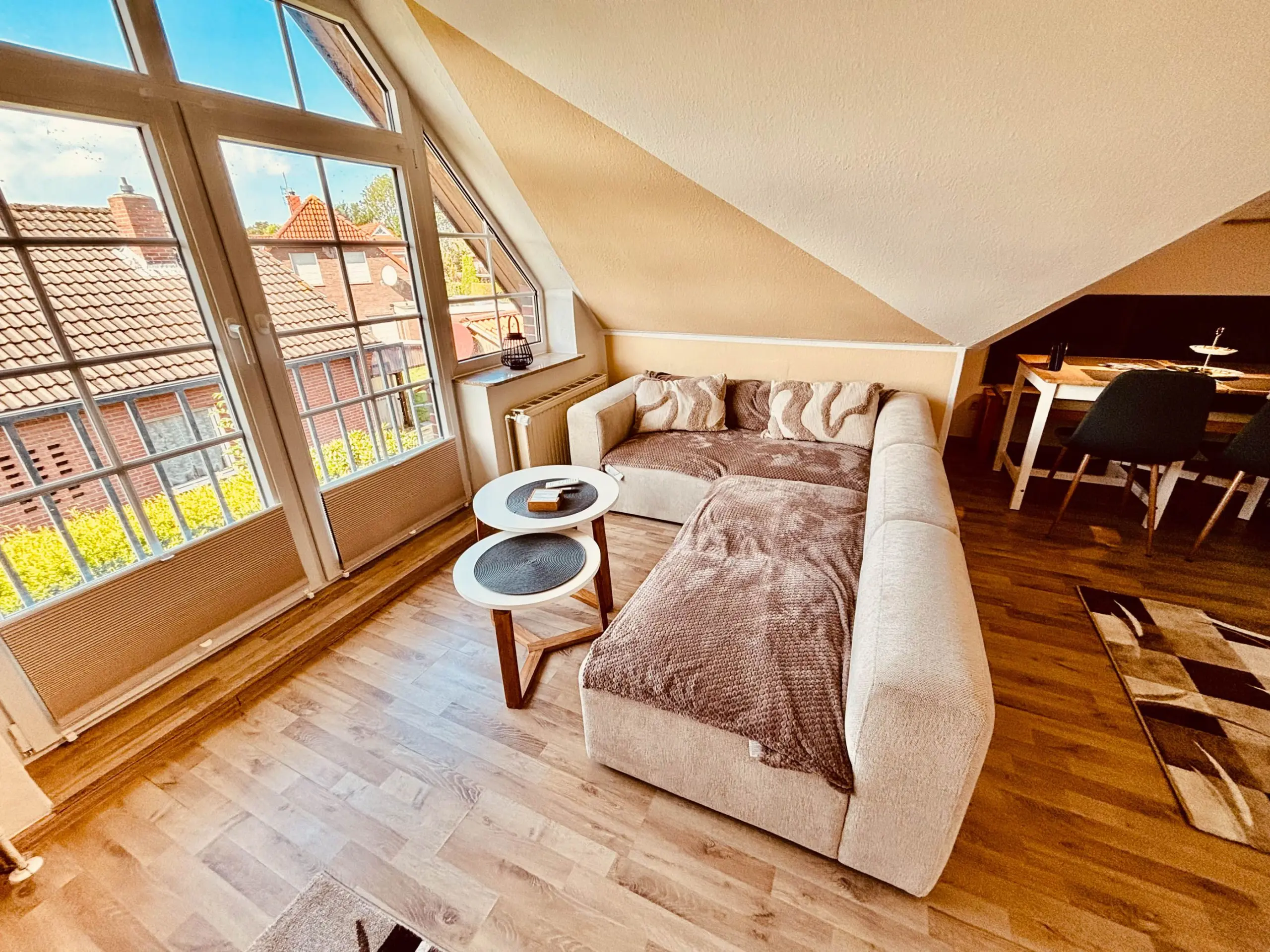 Living room with a tan sectional sofa, two round nesting coffee tables, and large angled windows letting in bright daylight. A dining area is in the back right.
