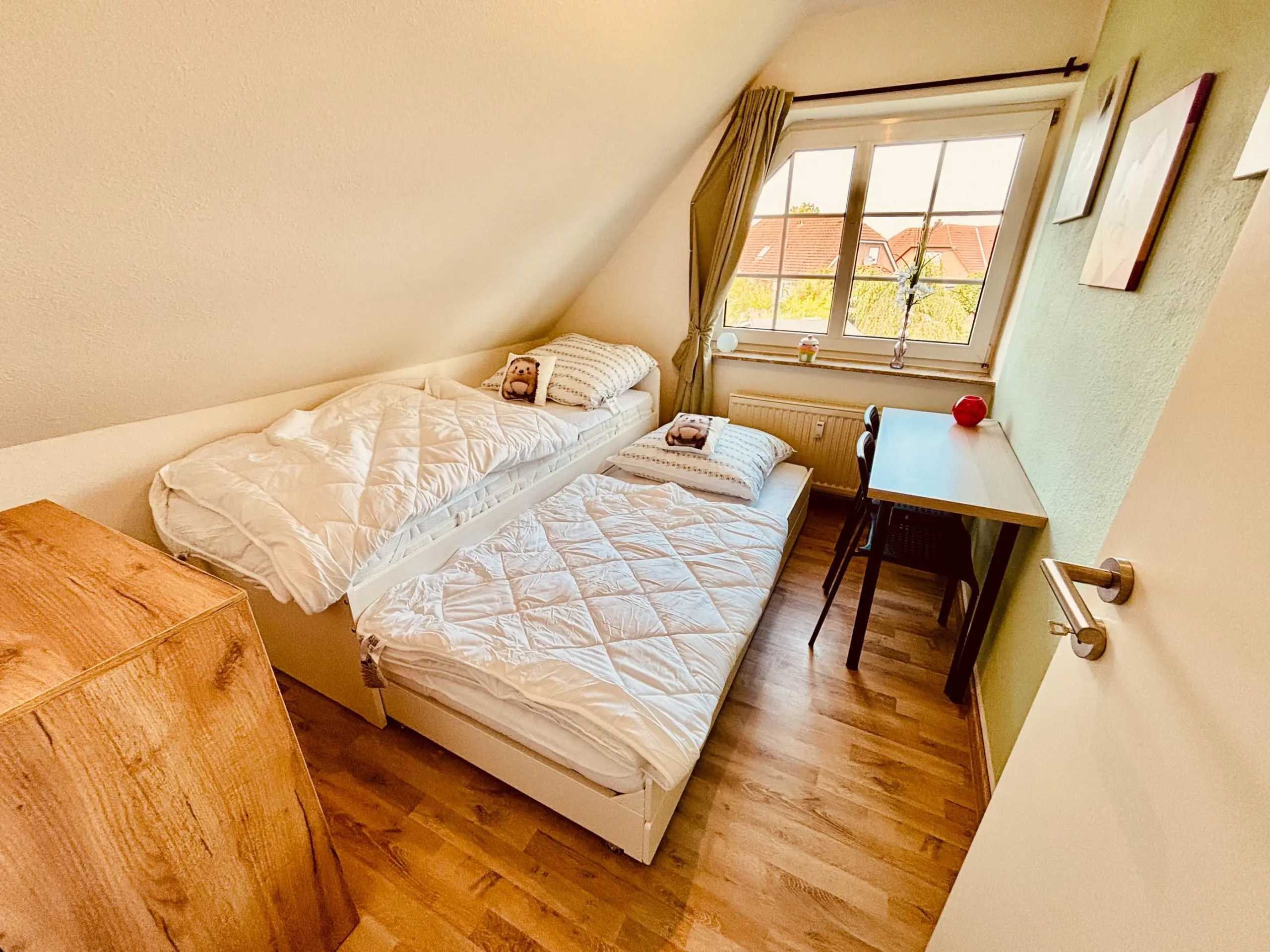 Twin beds with white quilts in a small attic bedroom, large window showing red-tiled rooftops.