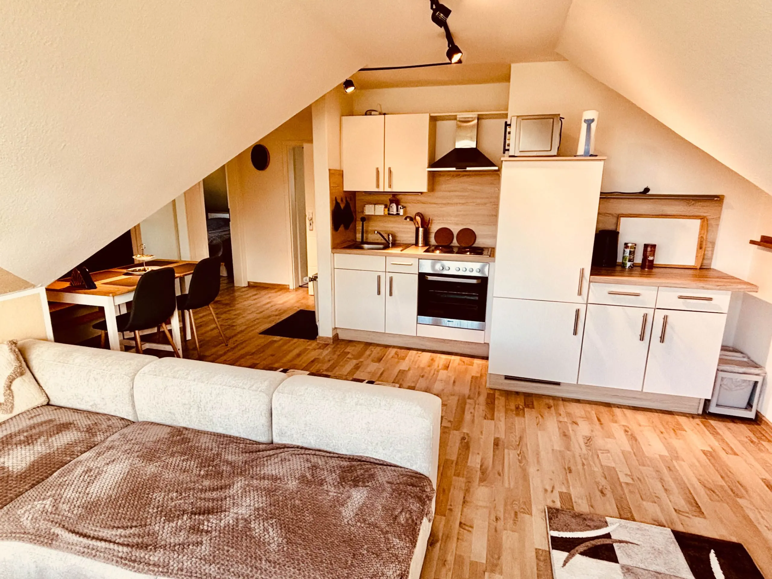 Open-plan living area with a kitchenette under a sloped ceiling: white cabinets, oven, sink, and a wooden countertop; a dining table with black chairs nearby; beige sofa in foreground.