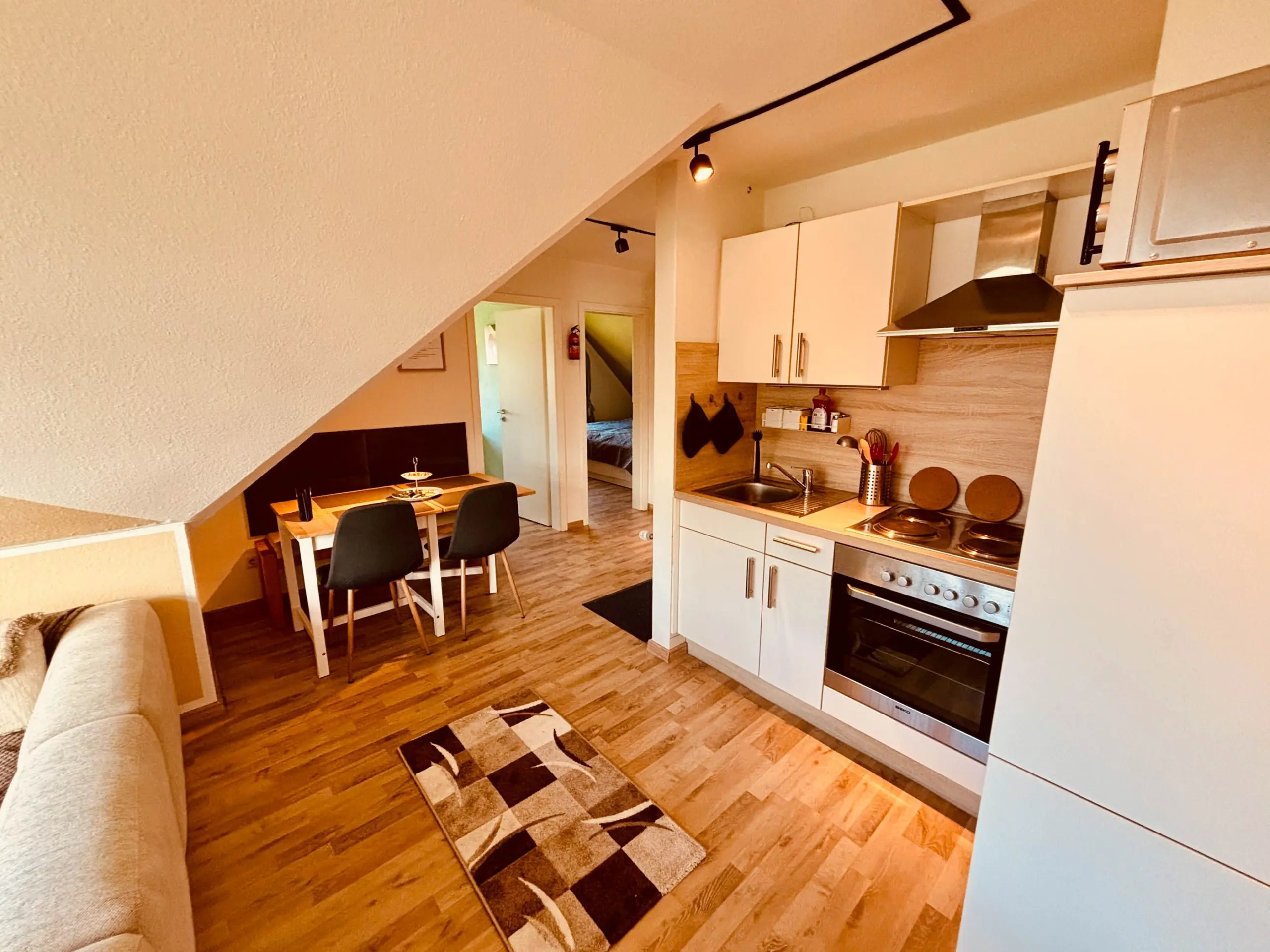 Open kitchen with white cabinets, stainless steel oven, sink, and a wooden dining table under a sloped ceiling.