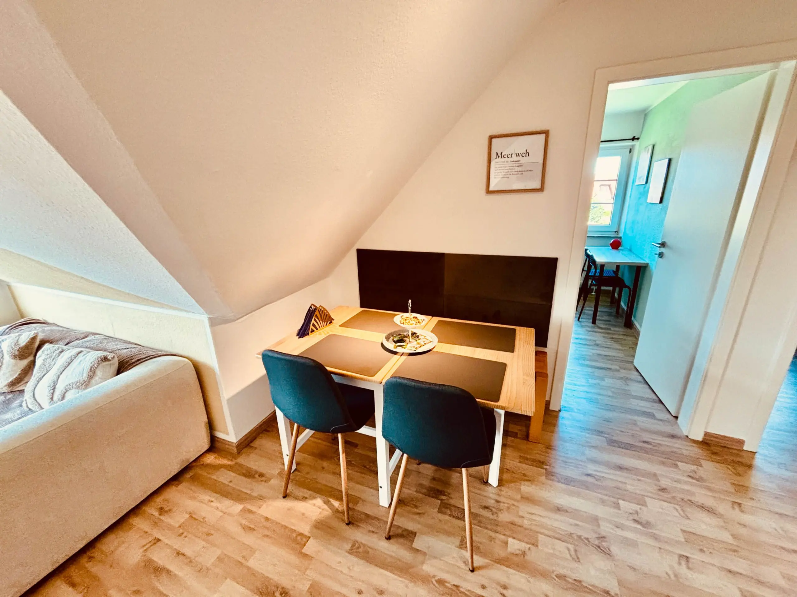 Small dining table with four teal chairs in a bright living area; sofa to the left and a framed wall plaque above the table, doorway to another room visible in the background.