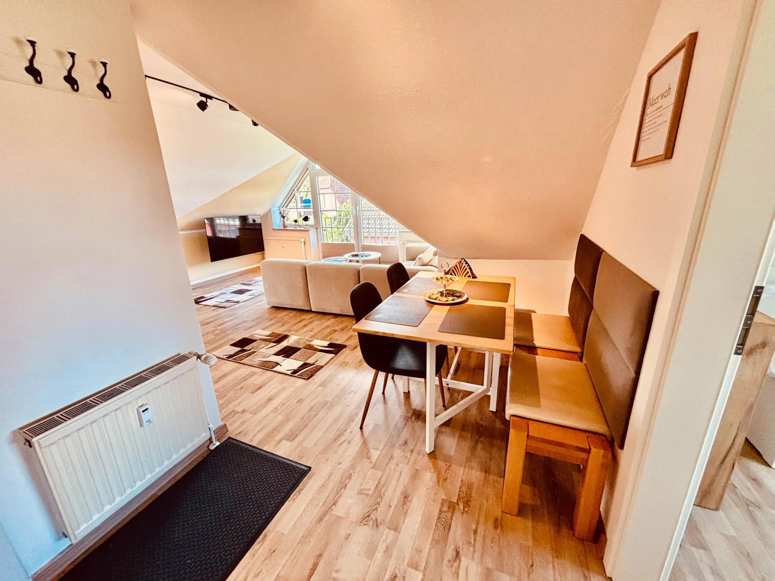 Open-plan living and dining area under a sloped ceiling, with a wooden dining table set for four and a sofa in the background by large windows.
