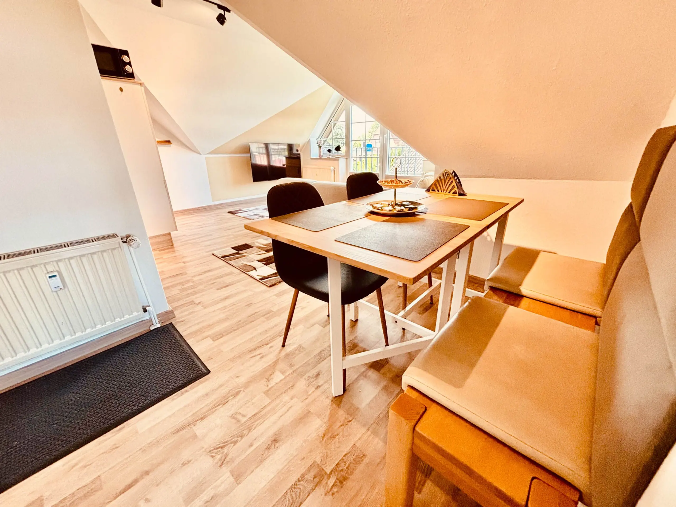 Attic dining area with a wooden table, black chairs, and a beige sofa under slanted white ceilings; window view outside left of center.
