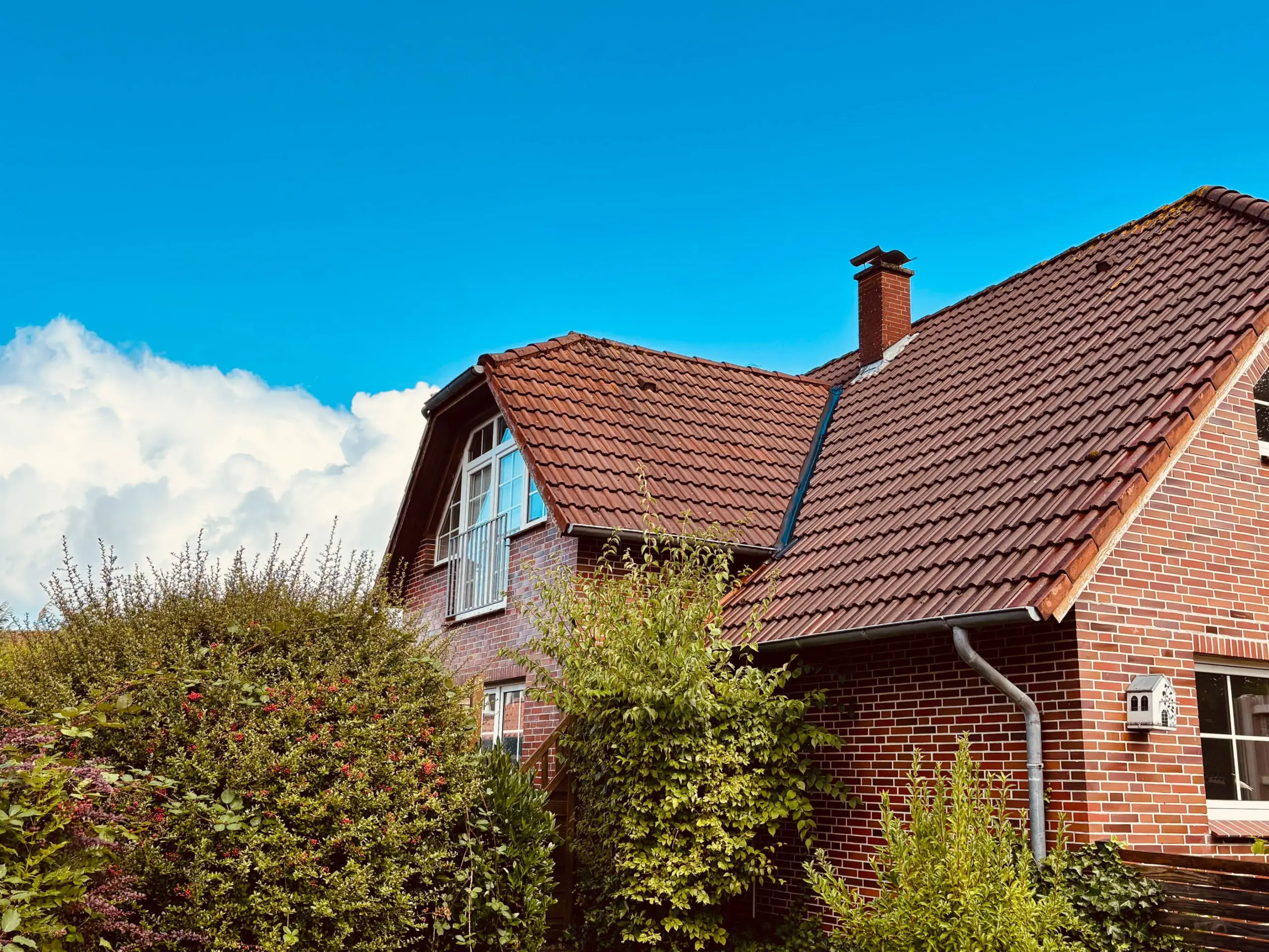 Brick house with a red tile roof and white-framed dormer windows under a bright blue sky. Tall brick chimney visible.