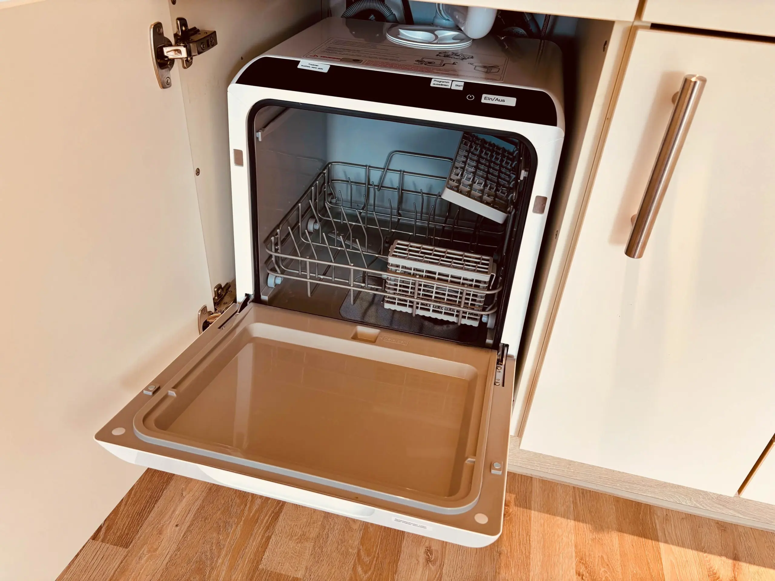 Open dishwasher drawer with racks pulled out, showing interior and dish rack on a wooden floor cabinet.