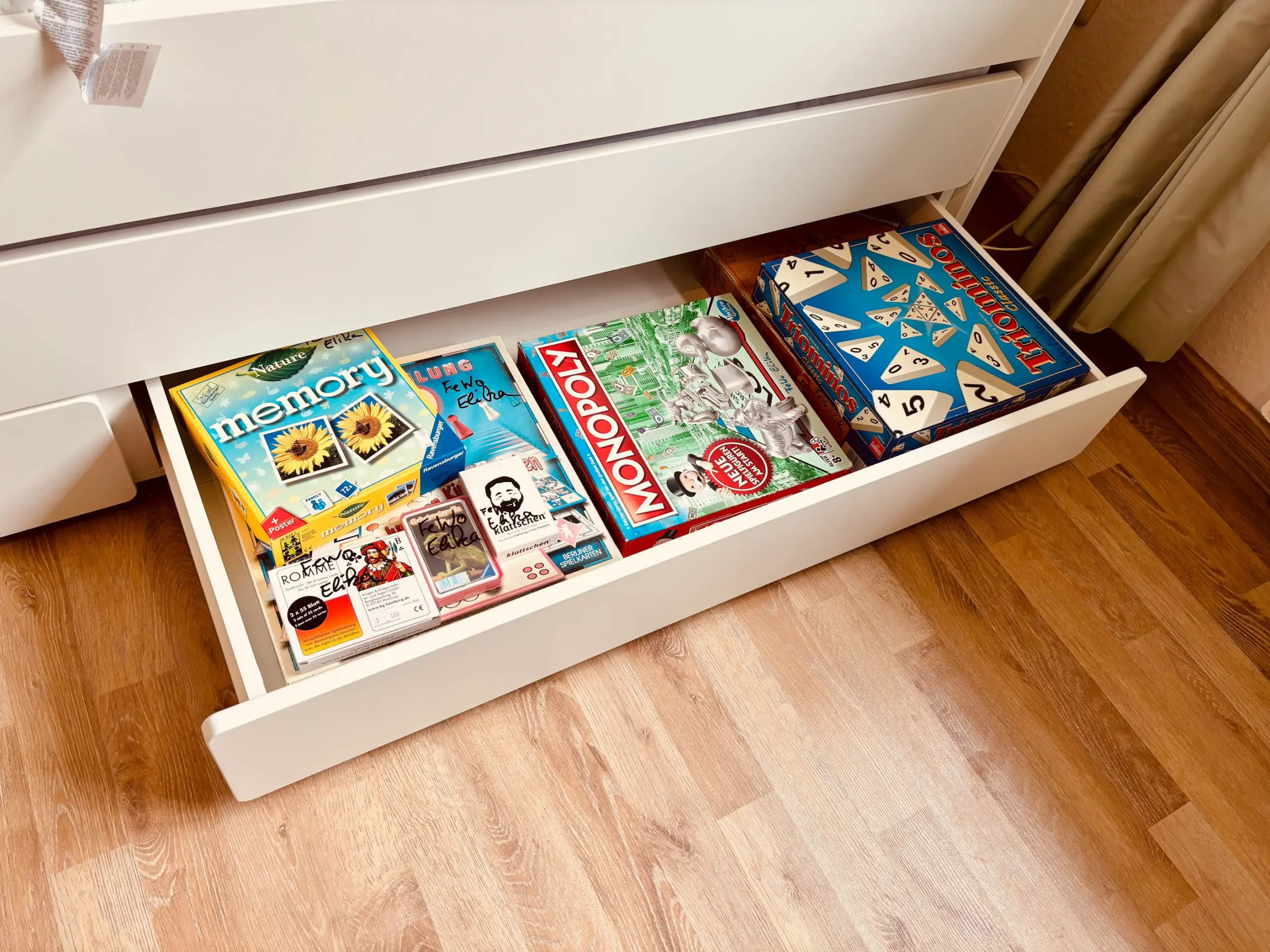 White dresser with the bottom drawer pulled out, showing assorted board games like Monopoly and Memory inside on a wooden floor.