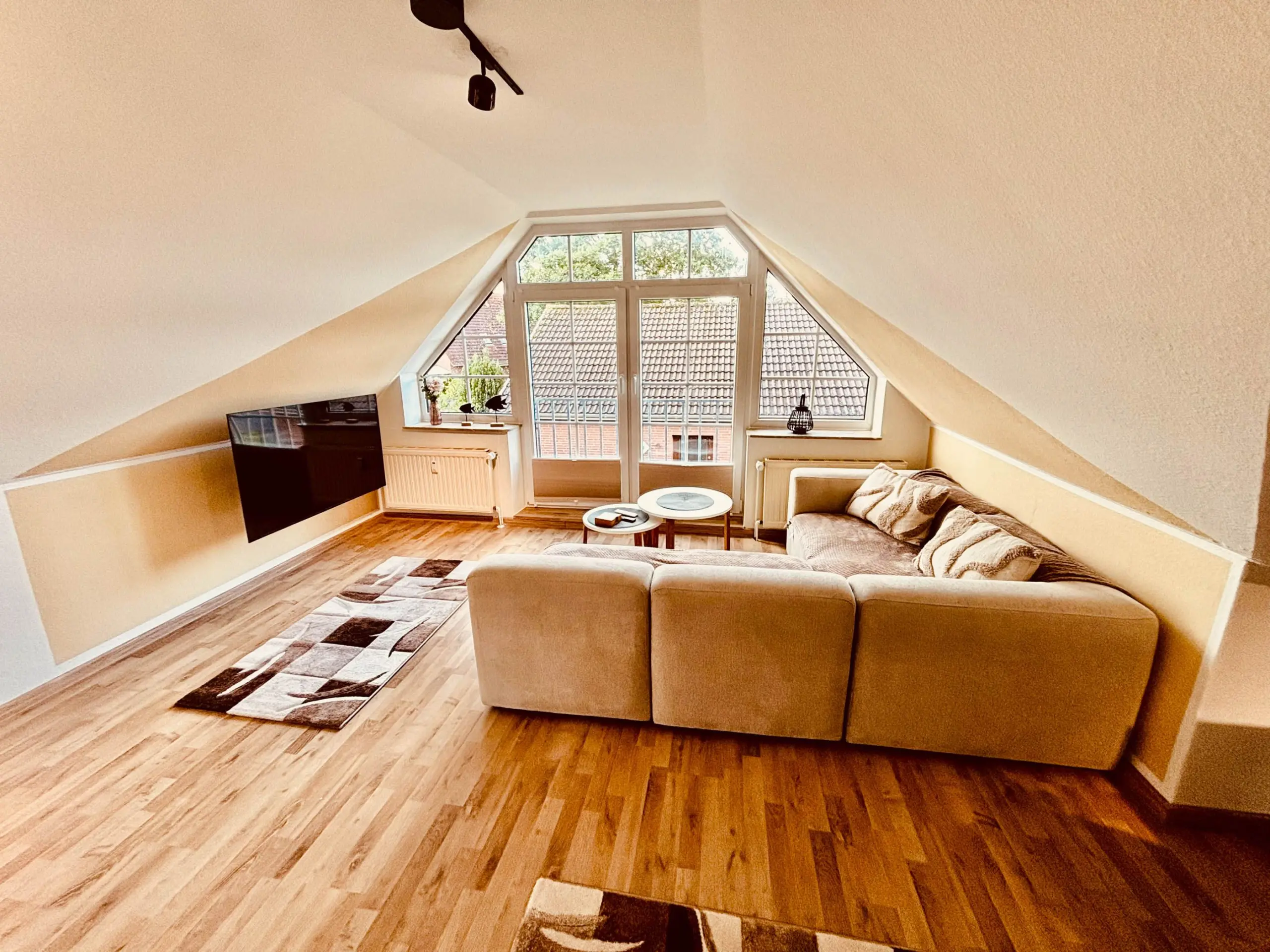 Bright attic living room with a tan sectional sofa facing a large triangular window, wooden floors, and a geometric rug.